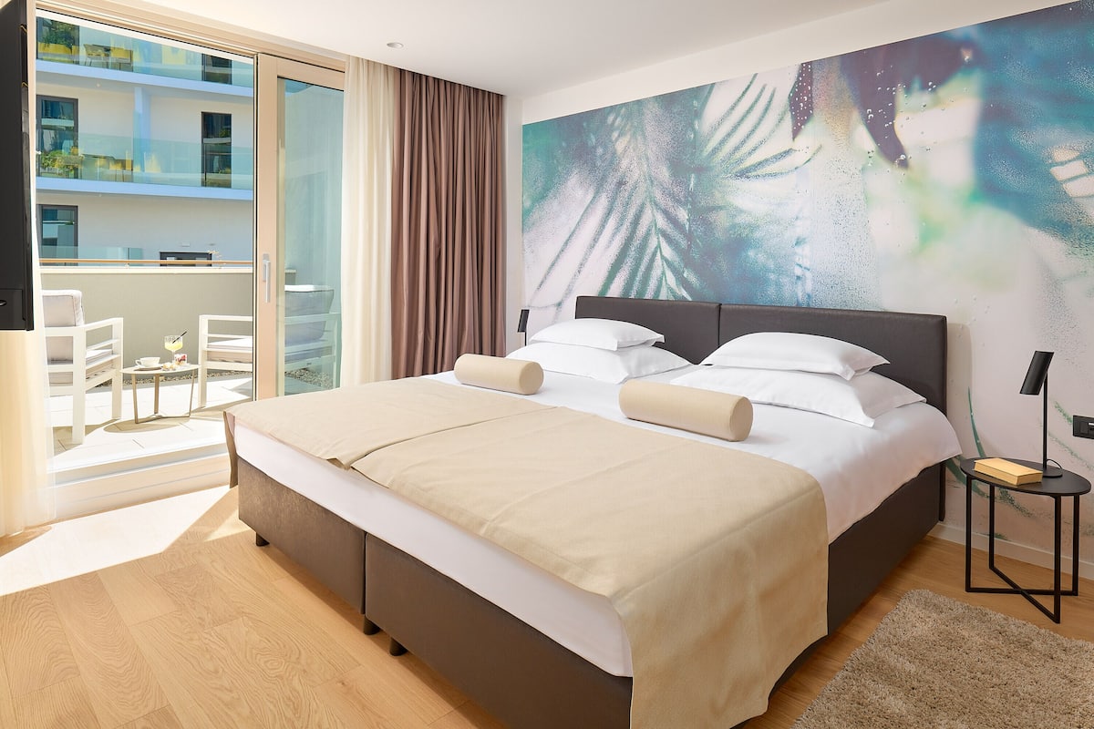 Ein modernes Hotelzimmer mit einem großen Bett, einem Balkon mit Blick auf das Gebäude und einem großen, abstrakten Gemälde an der Wand.