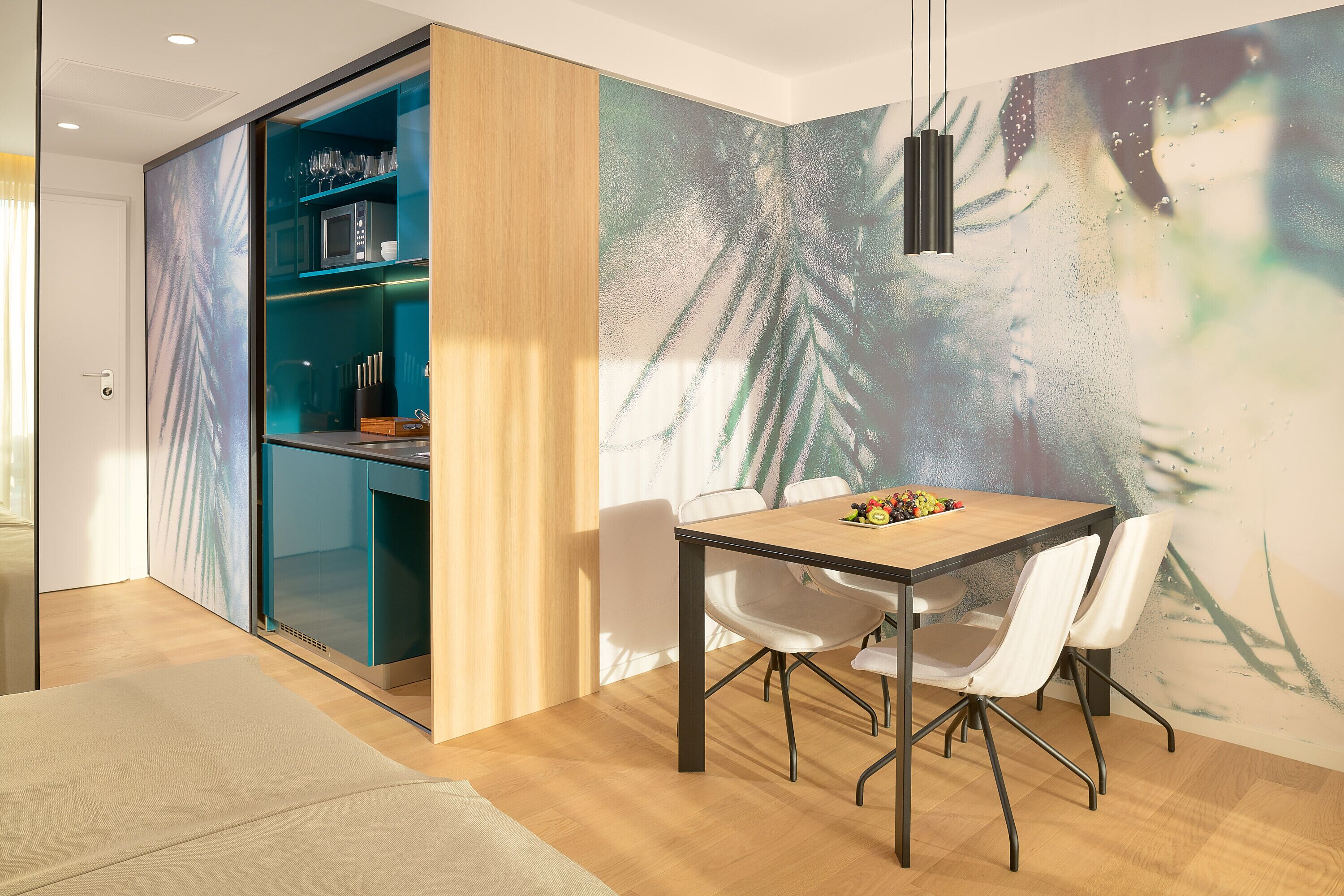 Een eetgedeelte met een tafel en stoelen, een keuken met blauwe kastjes en een muur met een bladmotief.