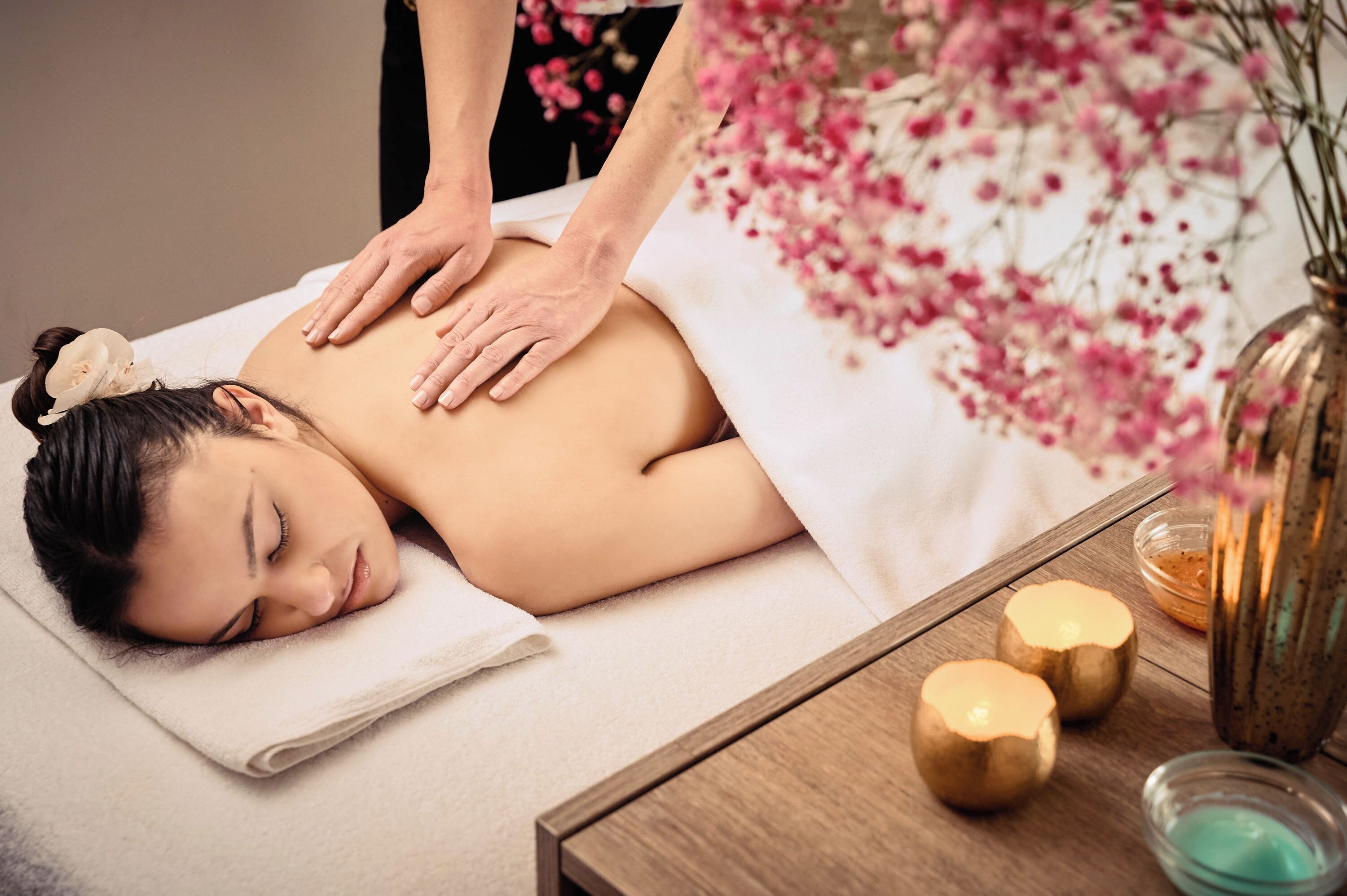 A person receiving a massage on a bed with a towel, a wooden table with candles, and a vase with flowers.