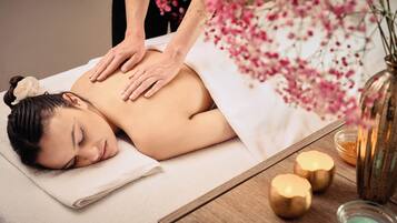 A person receiving a massage on a bed with a towel, a wooden table with candles, and a vase with flowers.