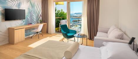 A hotel room with a bed, a desk, a chair, a TV, and a view of the sea.