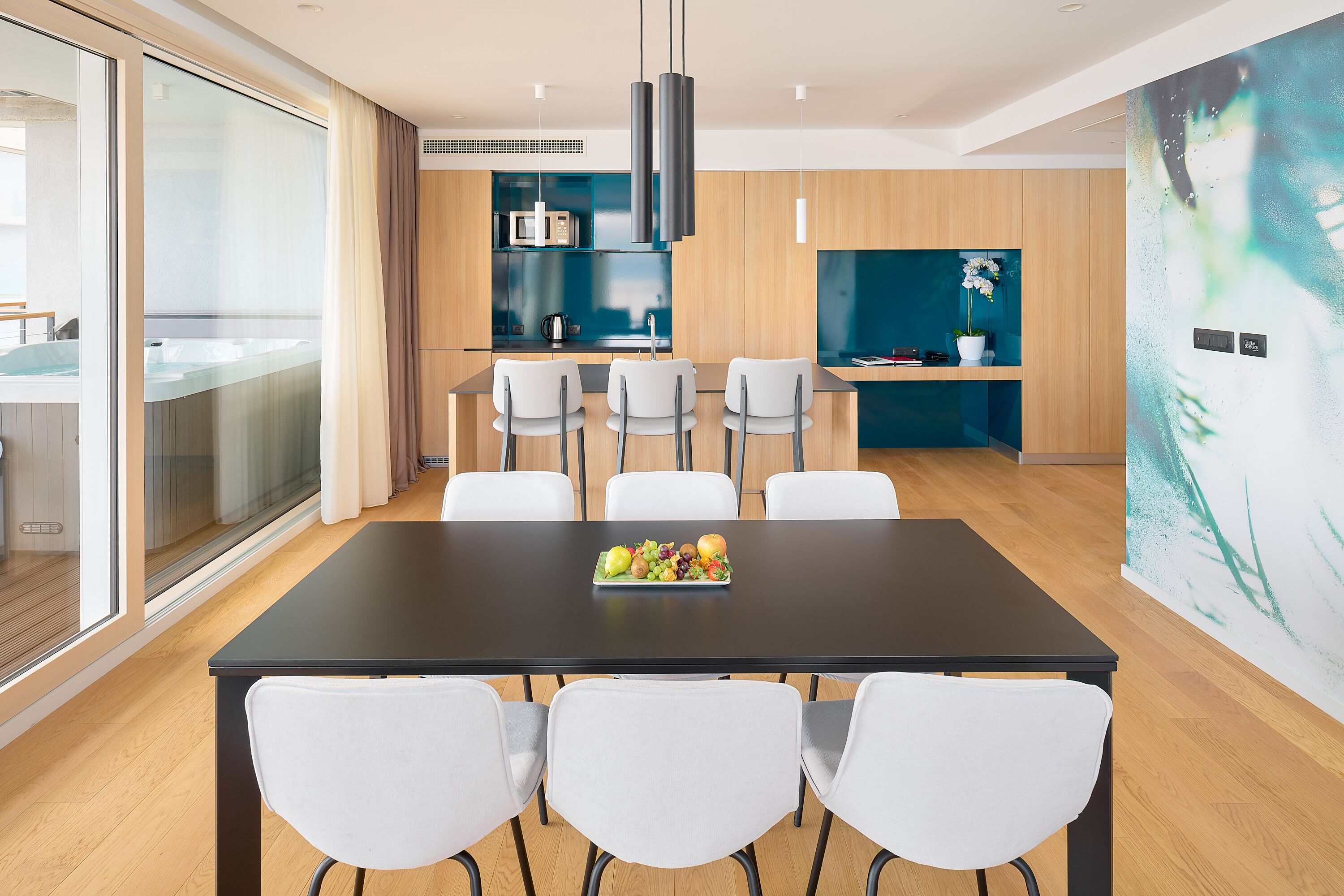 A modern dining area with a black table, white chairs, and a fruit bowl. There is a kitchen in the background with wooden cabinets and a blue accent wall.