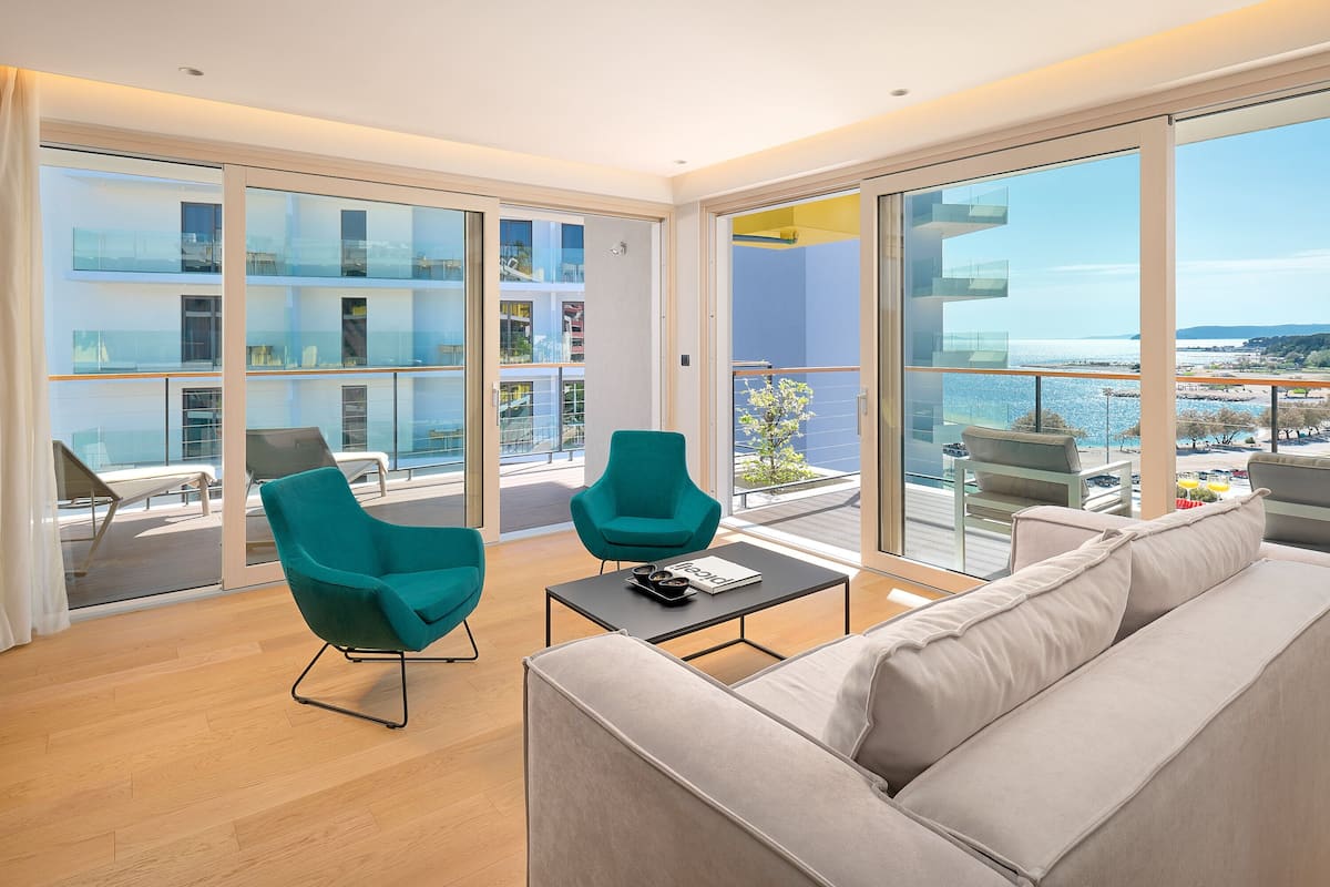 Ein modernes Wohnzimmer mit einer Couch, zwei türkisfarbenen Sesseln und einem Couchtisch. Große Glasschübe führen zu einem Balkon mit Blick auf das Meer und Gebäude.