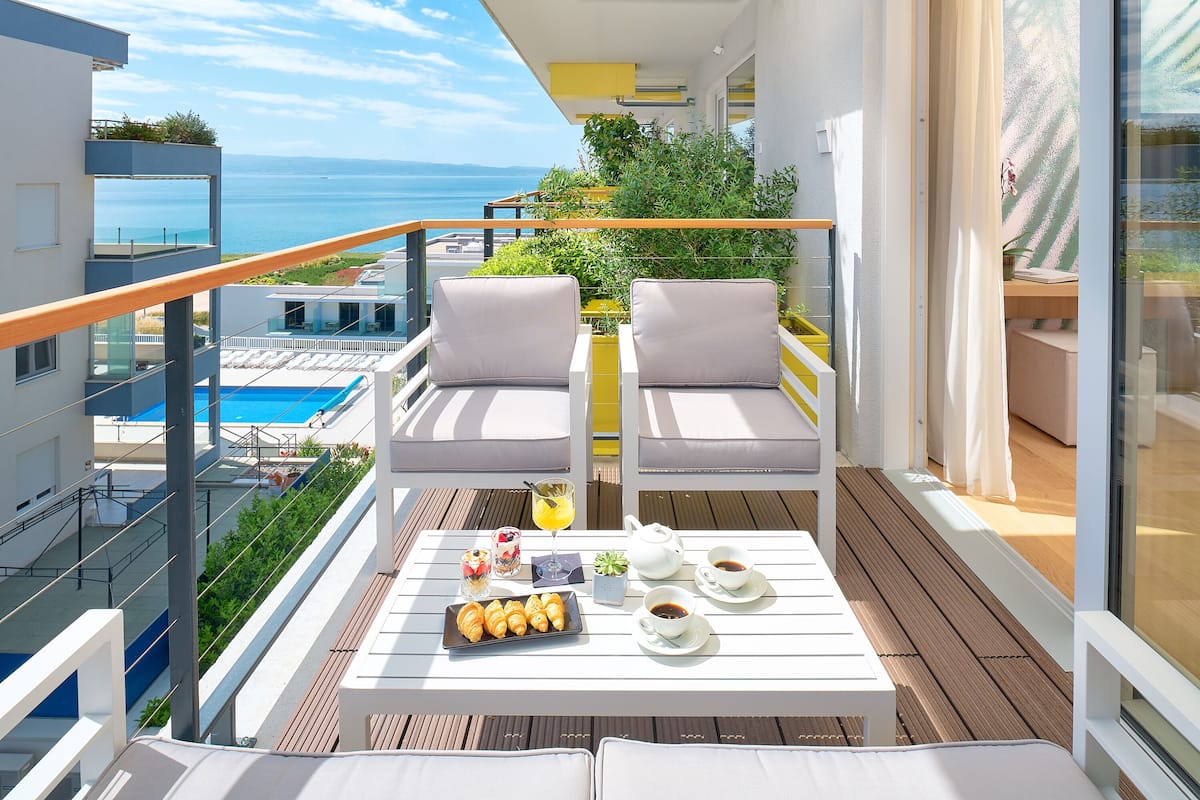 Ein Balkon mit zwei gepolsterten Stühlen, einem Tisch mit Frühstücksgebäck und Blick aufs Meer.