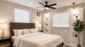 A bedroom with a bed, bedside tables, a ceiling fan, wall-mounted shelves with plants, and a large window with blinds.