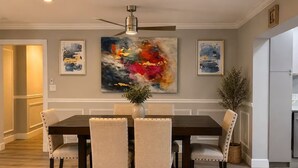 A dining area with a dark wooden table and four upholstered chairs, a ceiling fan, and abstract paintings on the wall.