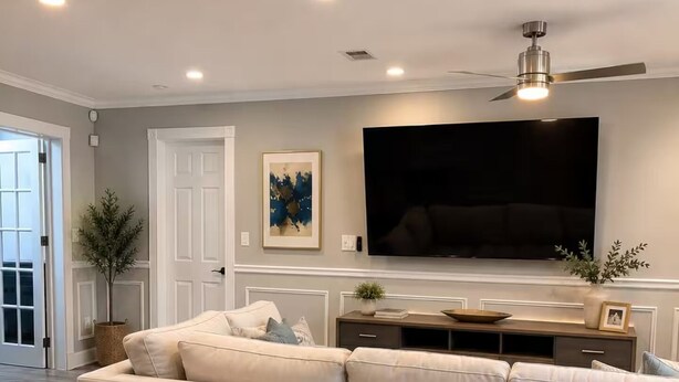 A modern living room with a sectional sofa, a flat-screen TV mounted on the wall, and a ceiling fan.