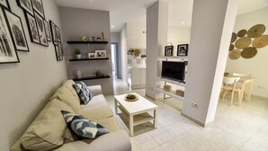 En moderne stue med en beige sofa, et hvidt sofabord og et fladskærms-tv. Reoler med dekorative genstande og et spiseområde med et hvidt bord og stole.