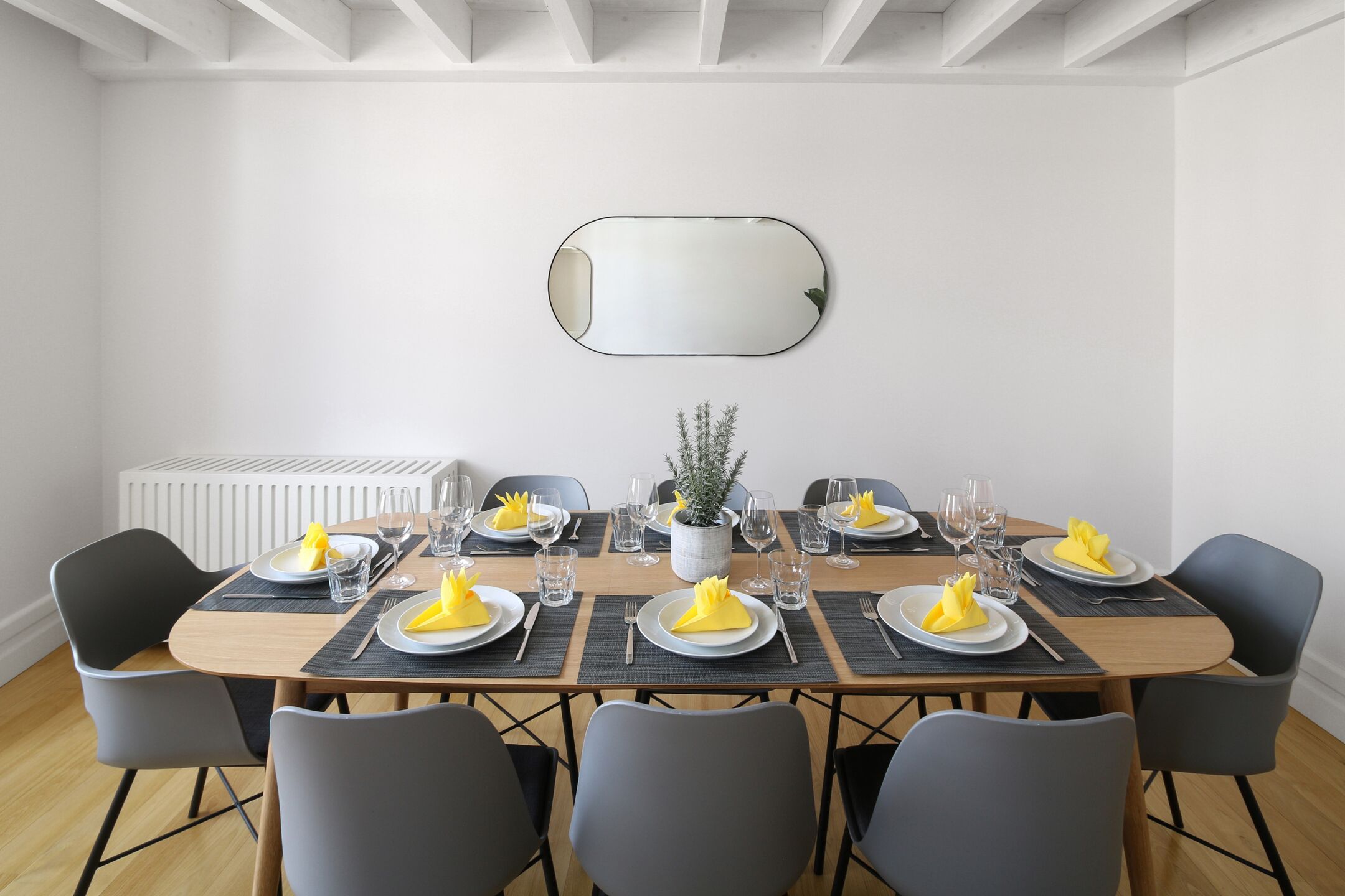 A dining room with a rectangular table set for six, surrounded by six chairs. A potted plant is centered on the table, and a mirror hangs on the wall above.