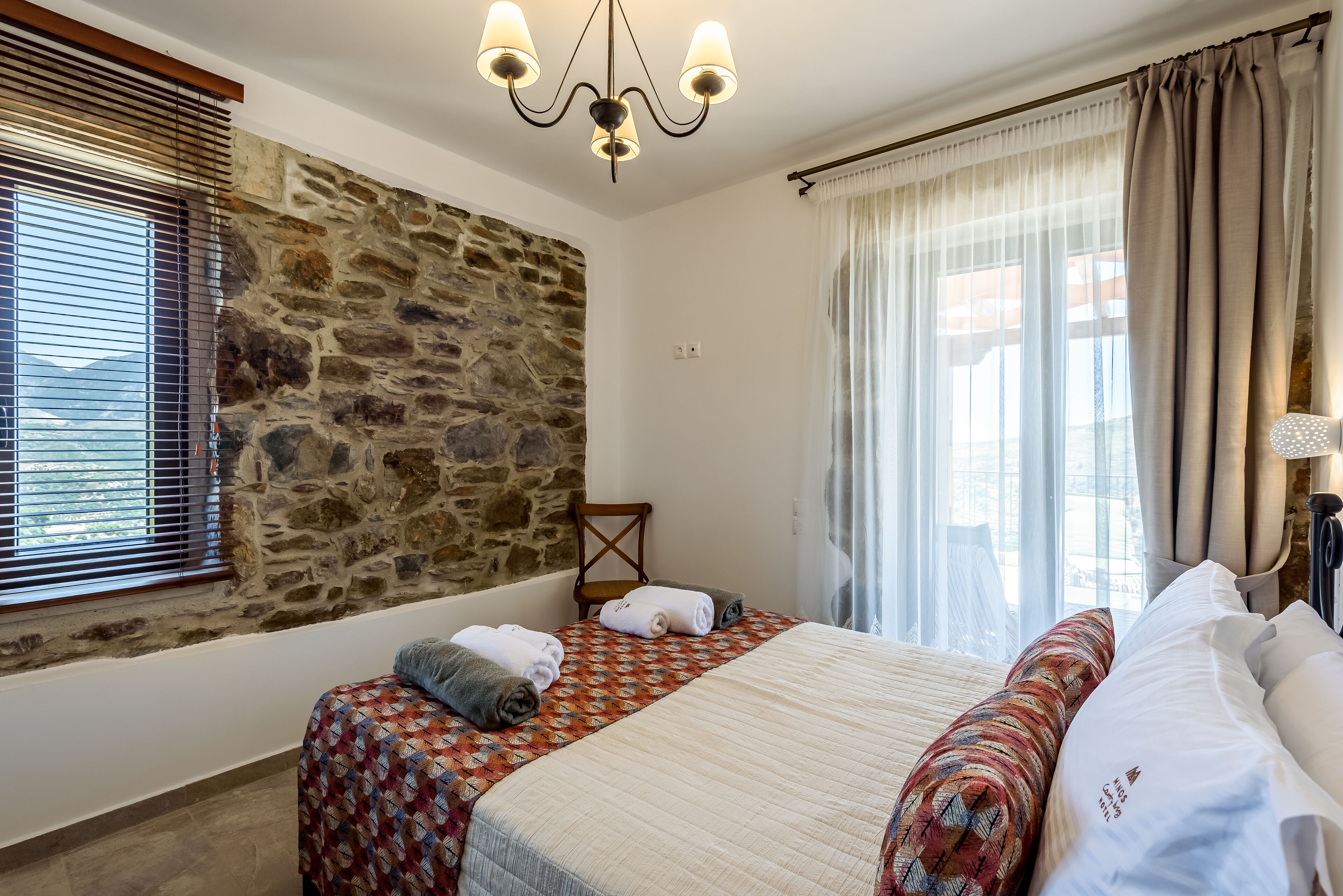 Ein Hotelzimmer mit Steinsandsteinwand, einem Bett mit gemustertem Überwurf, einem Chandelier und Blick durch transparente Vorhänge.