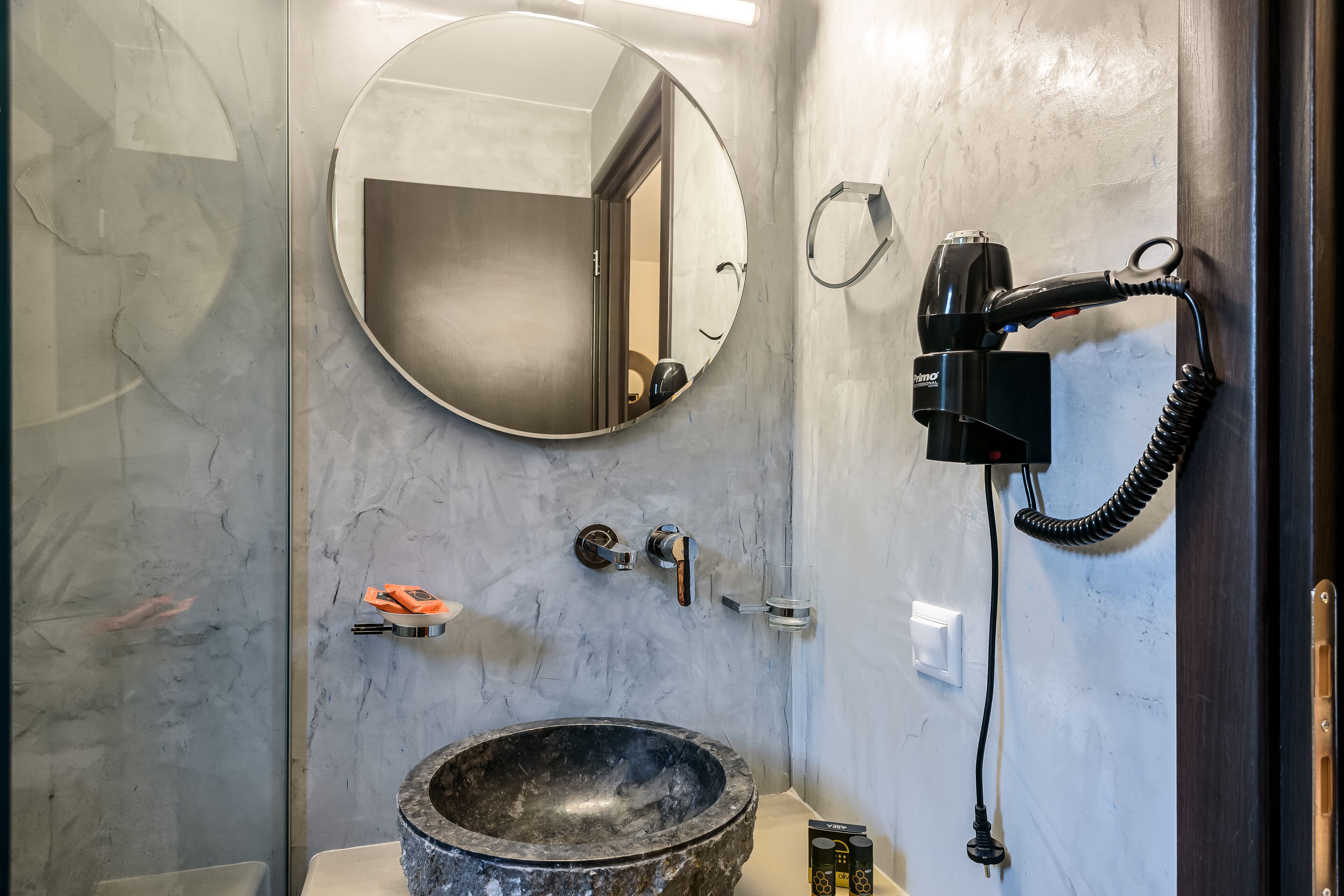 A bathroom with a round mirror, a sink with a stone basin, a hairdryer mounted on the wall, and a towel rack.