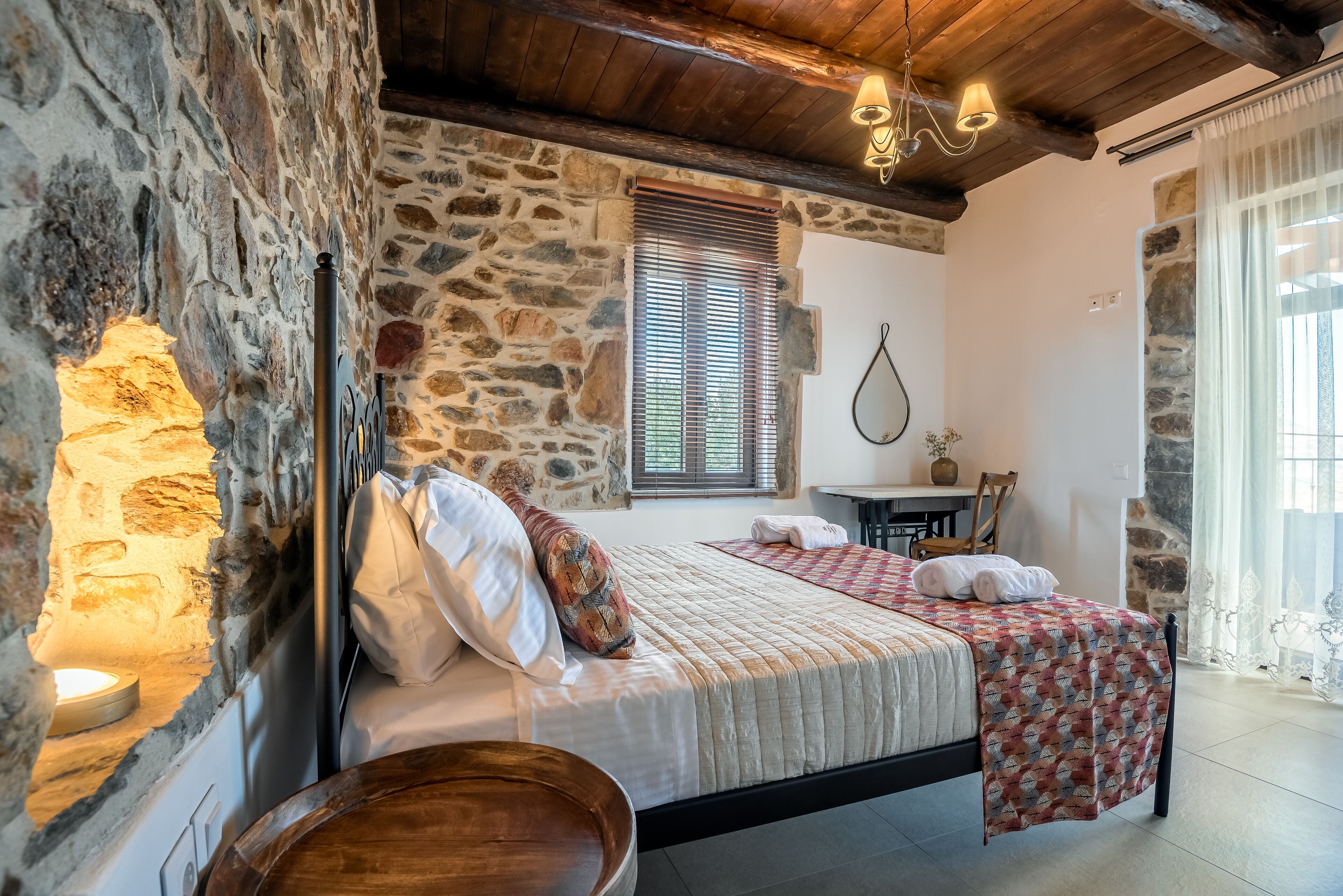 A bedroom with stone walls, a wooden bed, a small table, and a window with blinds.