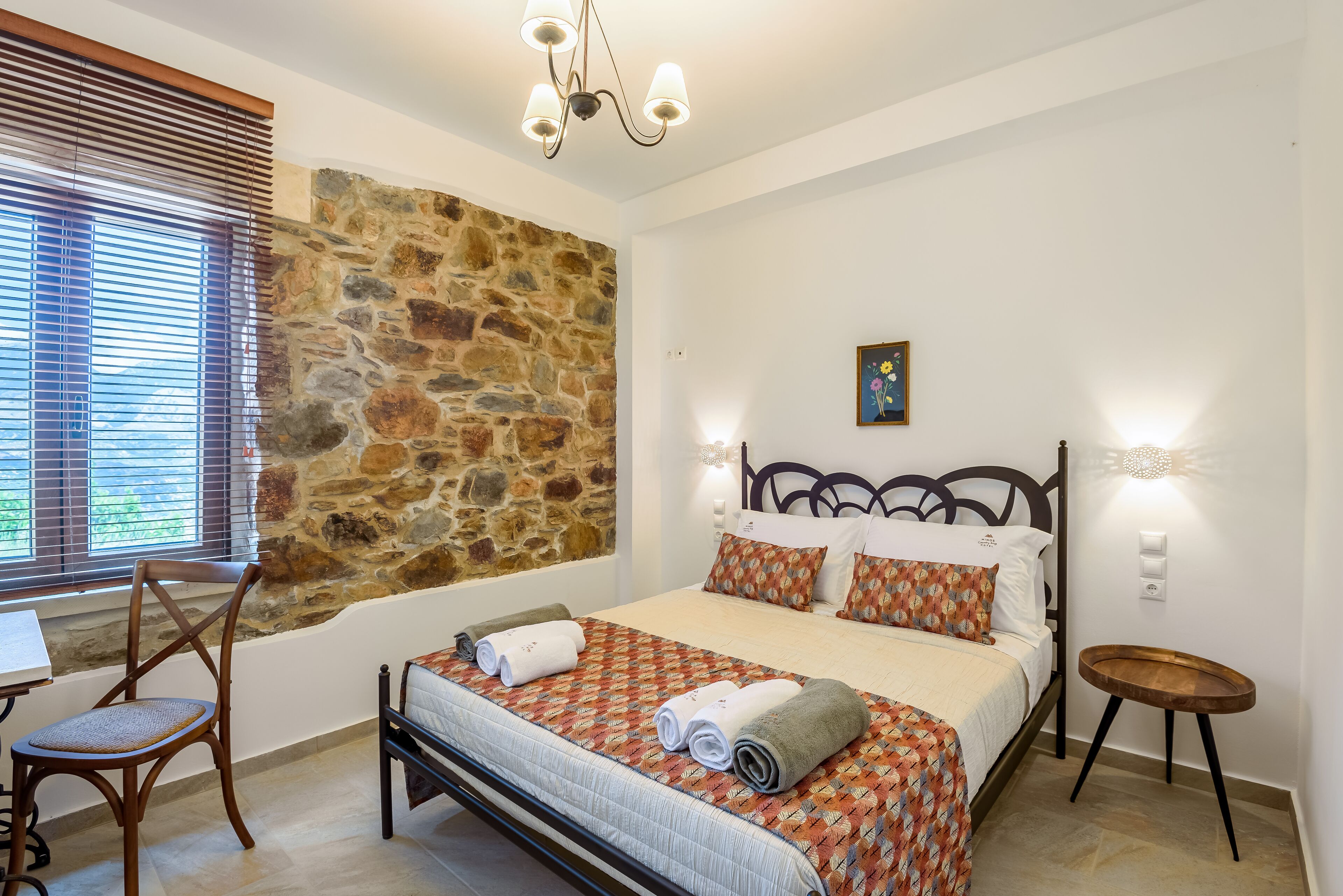 A bedroom with a stone wall, a bed with patterned bedding, a wooden chair, and a small table.