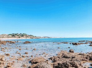 Une plage rocheuse aux eaux cristallines, avec une côte lointaine parsemée de bâtiments.