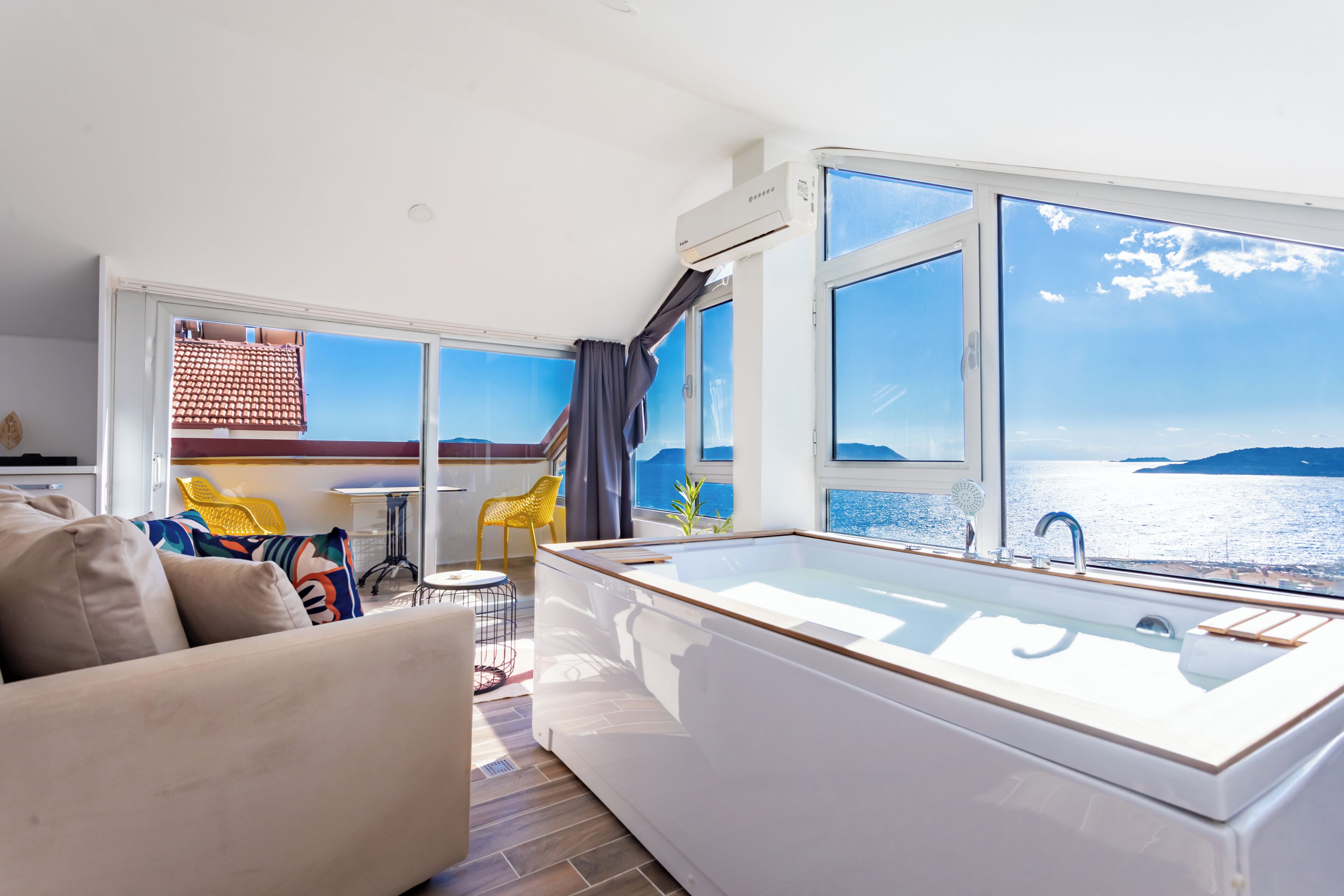 A modern room with a large window, a bathtub, a sofa, and a dining area with yellow chairs.