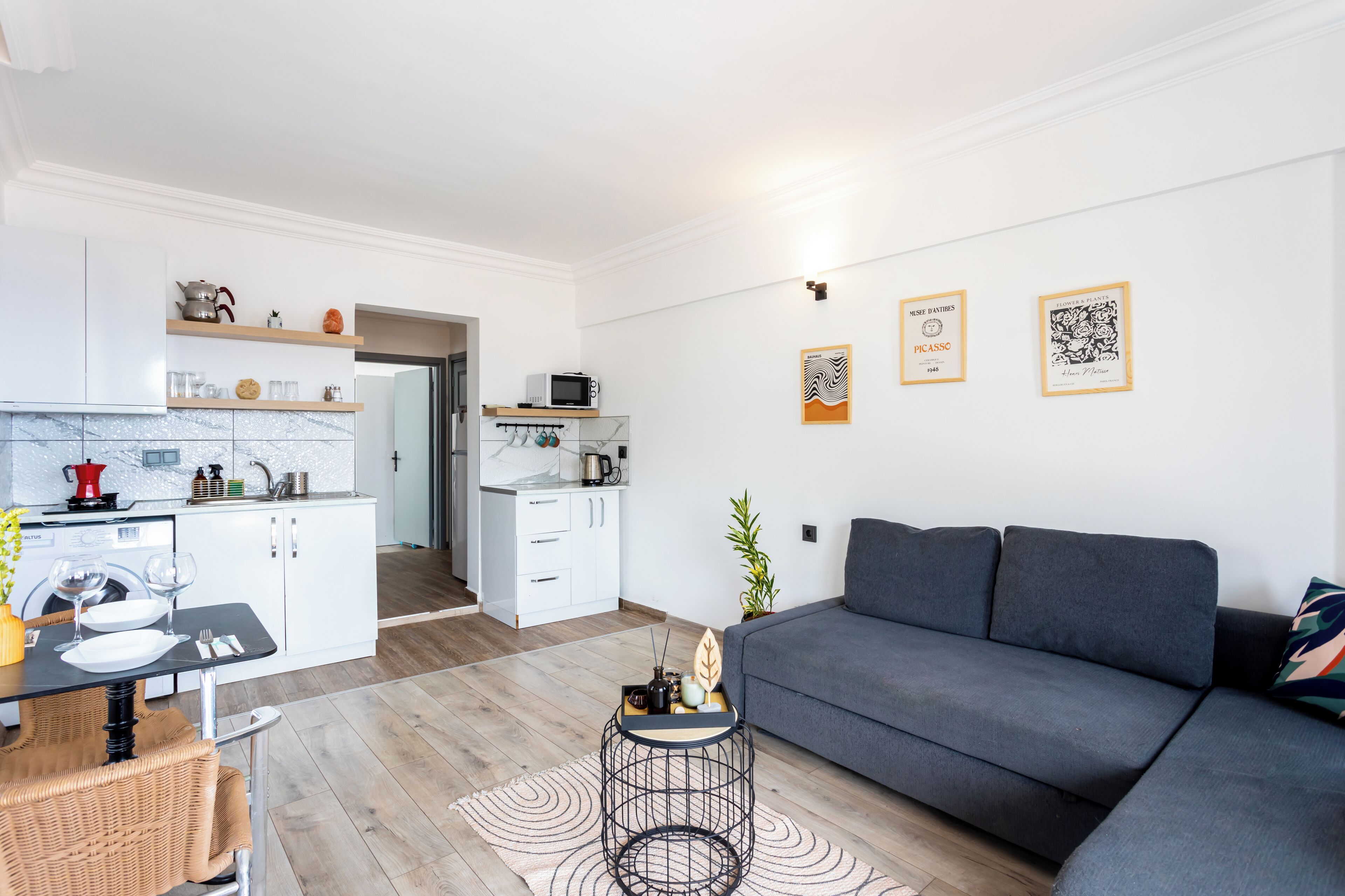 Ein modernes Wohnzimmer mit grauem Sofa, einem Esstisch mit Stühlen und einer Küchenzeile mit weißen Schränken und einer Mikrowelle.