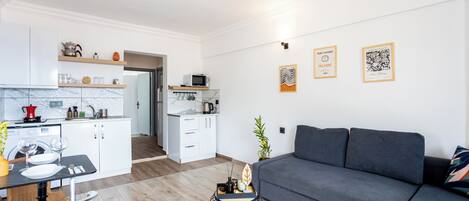 Ein modernes Wohnzimmer mit grauem Sofa, einem Esstisch mit Stühlen und einer Küchenzeile mit weißen Schränken und einer Mikrowelle.