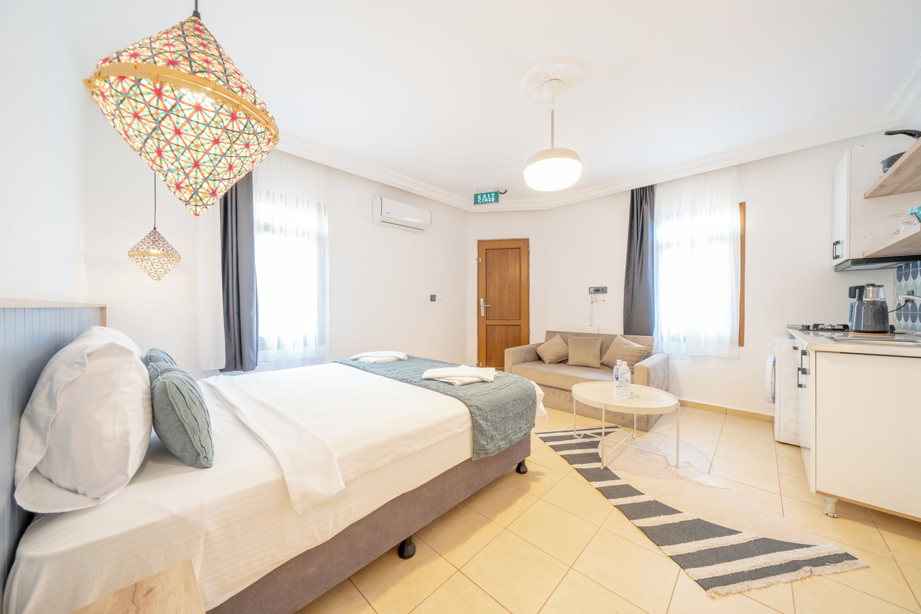 A modern hotel room with a bed, a sofa, a small round table, a kitchen area, and a decorative hanging light fixture.
