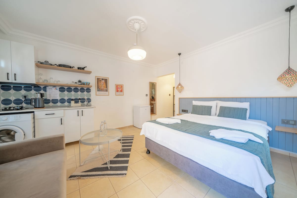 Een compacte hotelkamer met kitchenette, een bed met blauwe hoofdeinde en een klein zithoekje.