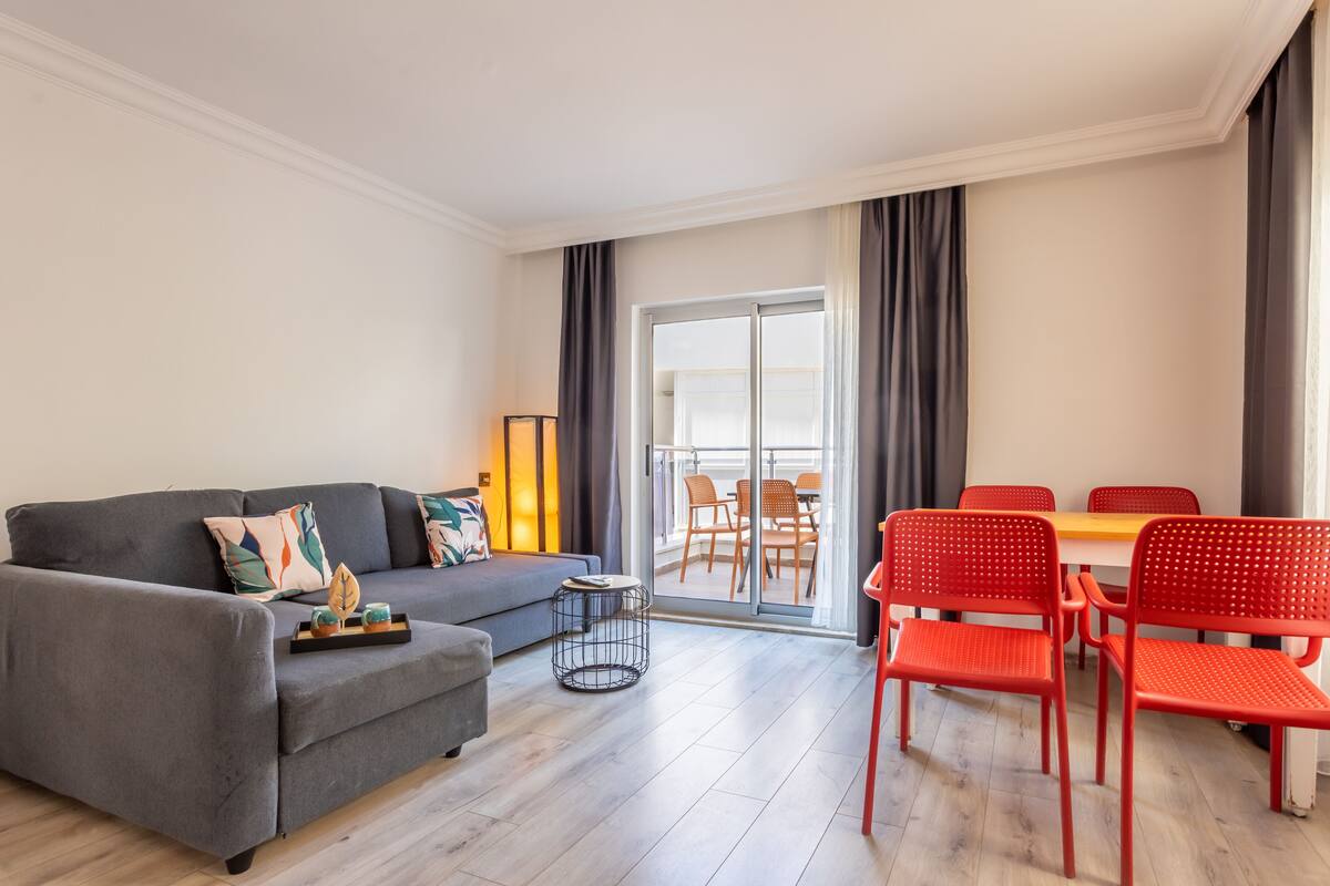 Een moderne woonkamer met een grijze bank, rode stoelen, een kleine ronde tafel en een balkon met een eethoek.