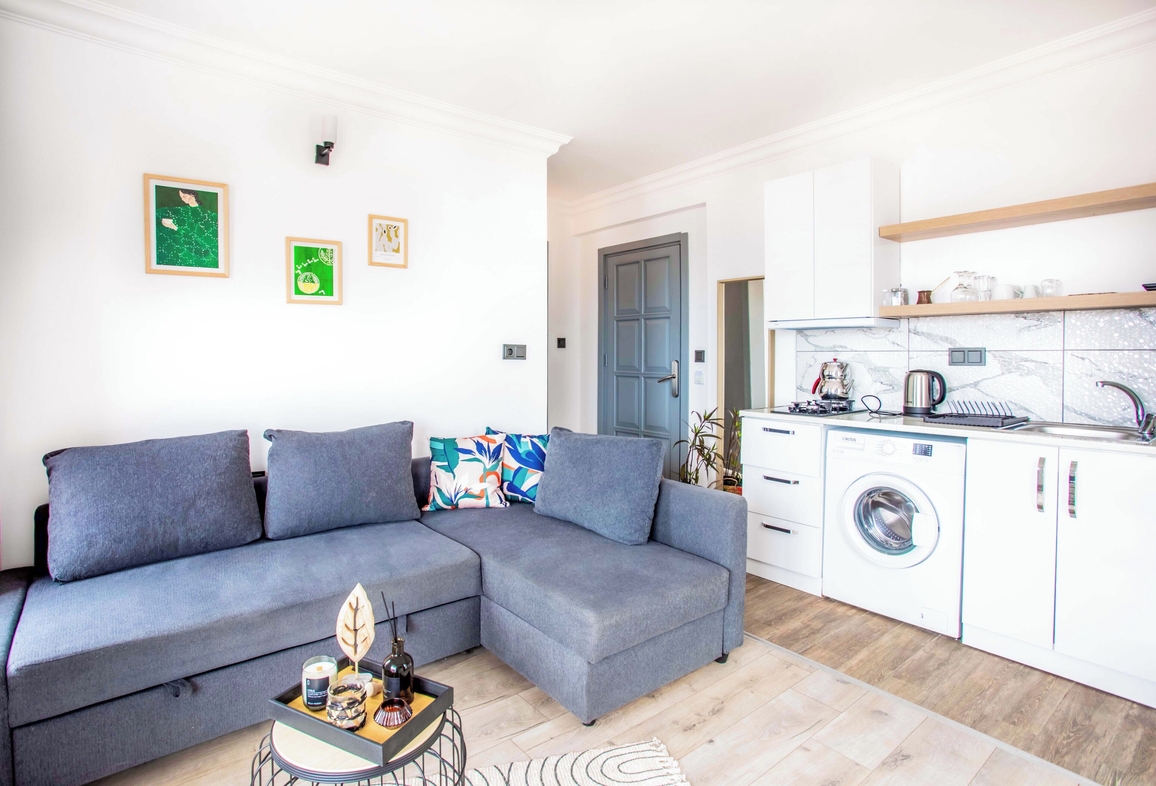 A modern living room with a grey sectional sofa, a small coffee table, and a washing machine in the adjacent kitchen area.