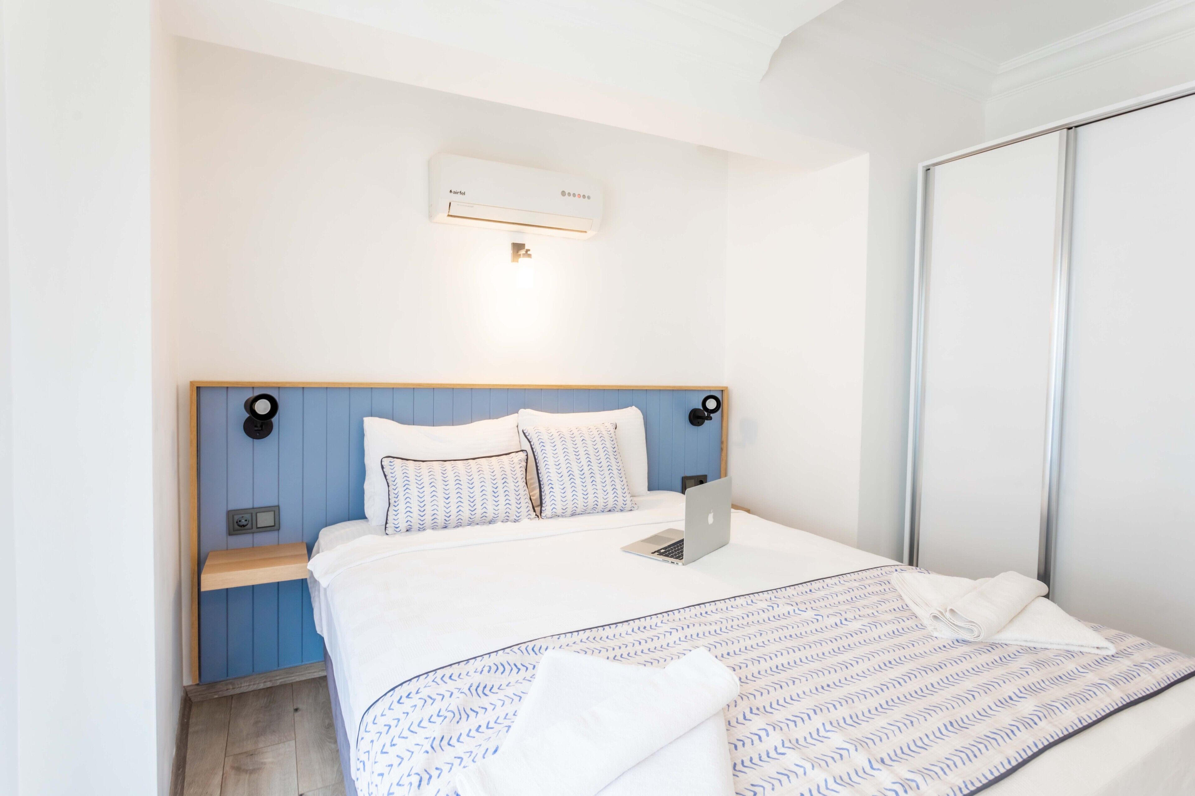 A neatly made bed with a laptop, a bedside table, and a wall-mounted air conditioner.