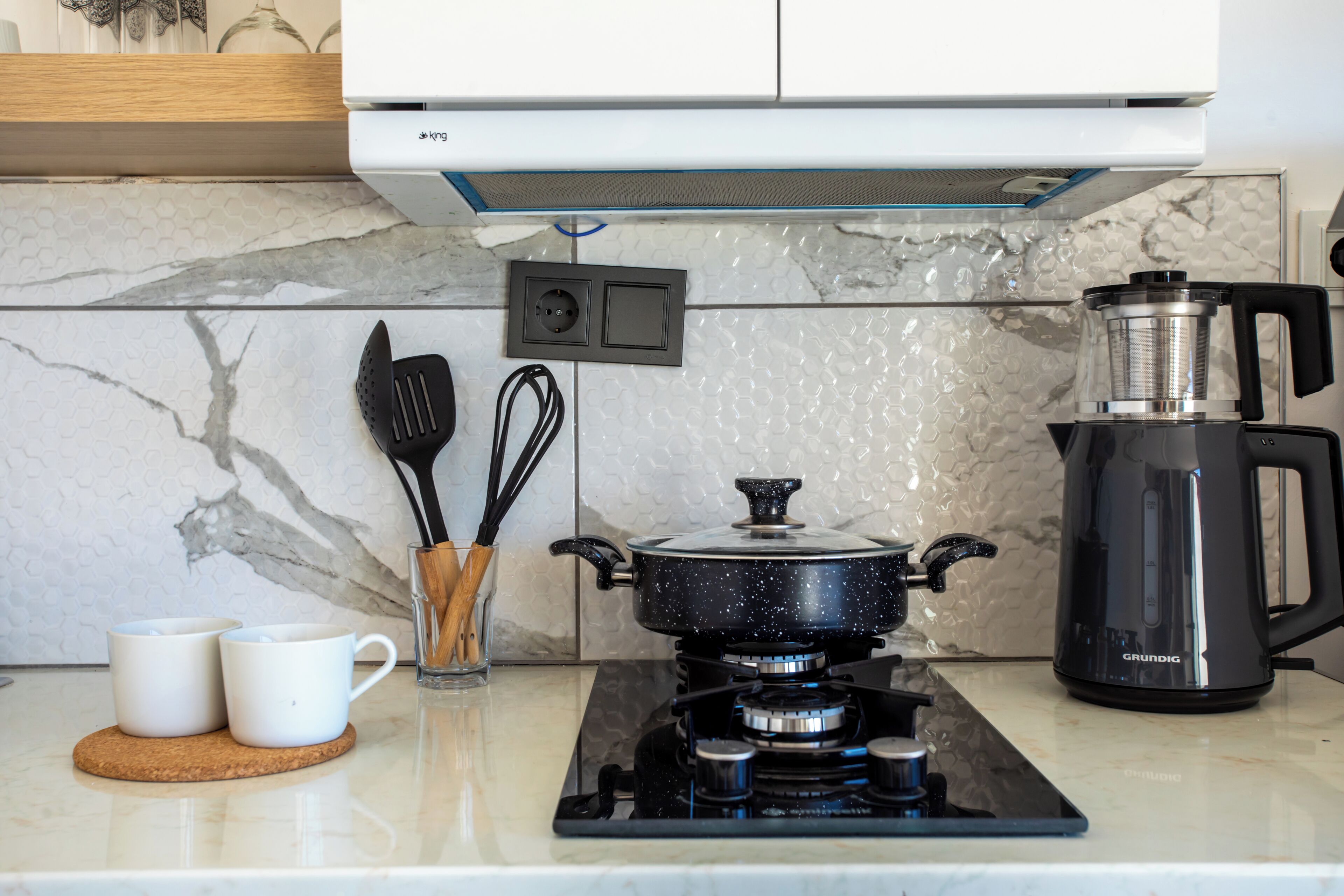 A kitchen with a gas stove, a coffee maker, and two white mugs on a marble countertop.