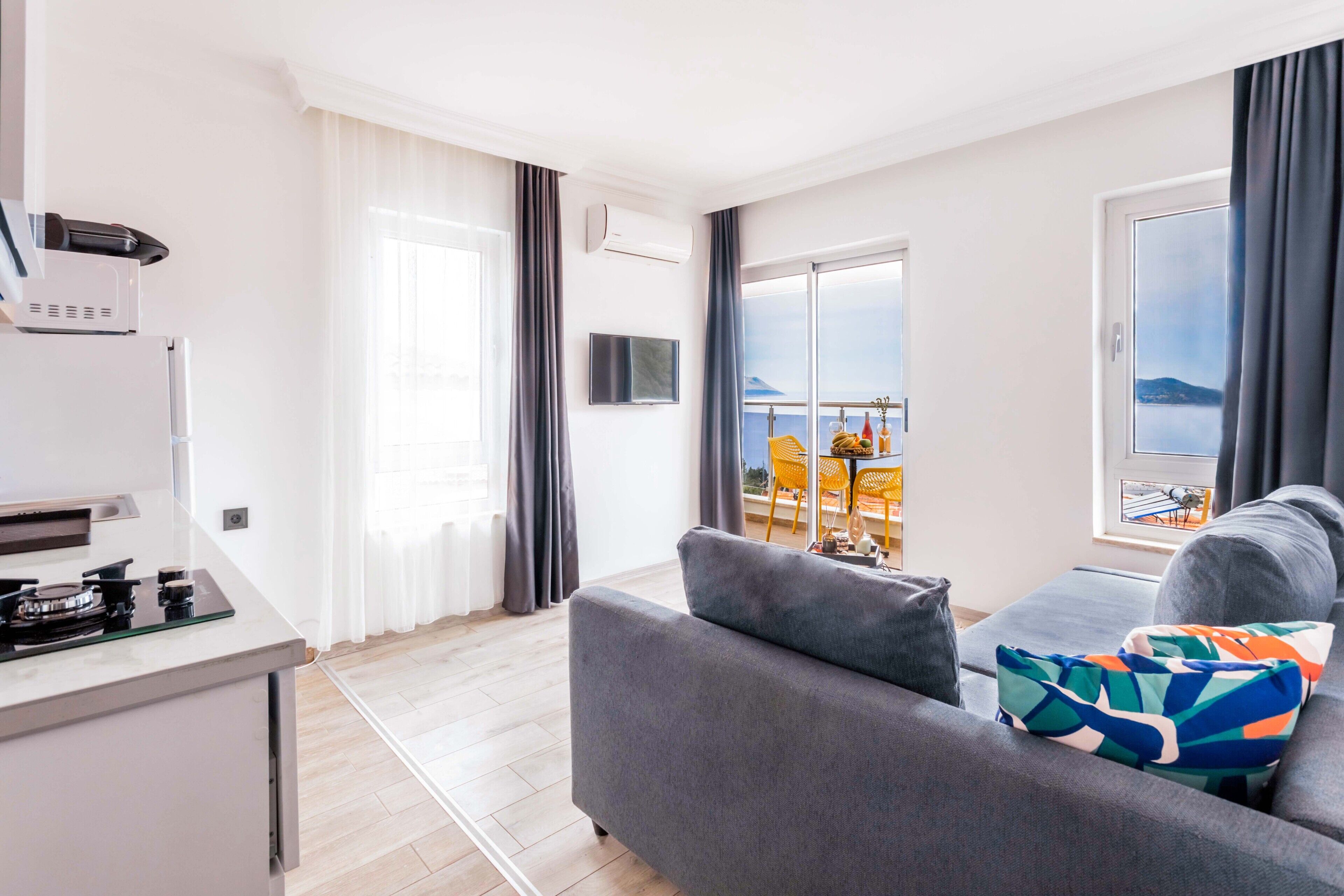 A modern living room with a grey sofa, a small kitchen area with a stove, a flat-screen TV, and a balcony with a table and chairs.