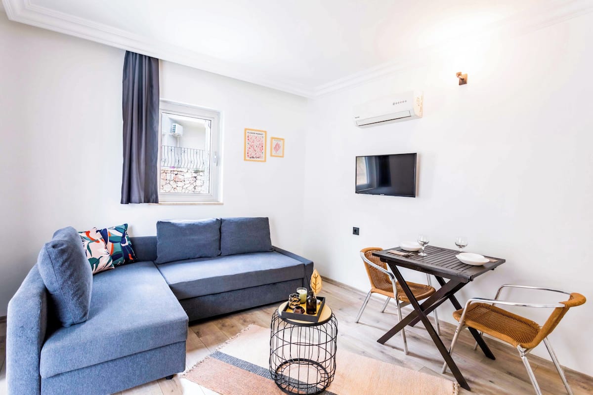 Een moderne woonkamer met een grijze modulaire bank, een ronde salontafel, een eettafel met stoelen, een flatscreen televisie en een raam met gordijnen.