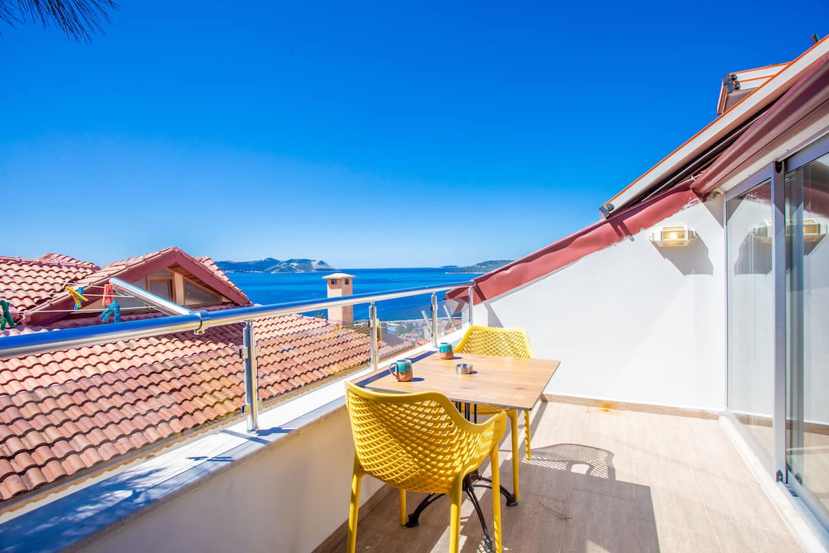 Een balkon met een tafel en gele stoelen, met uitzicht op een kustgebied met rode daken en de zee.