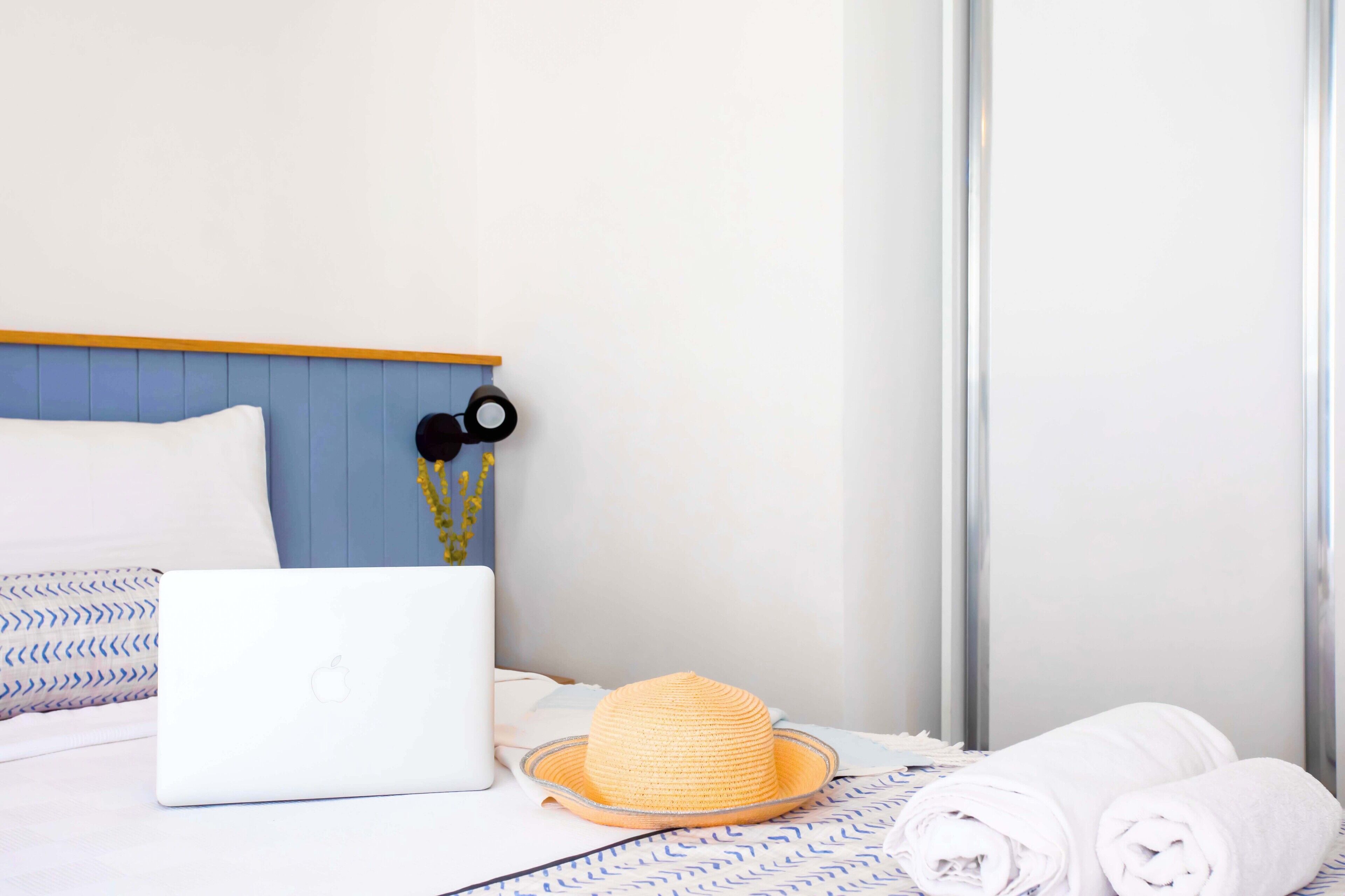 A neatly made bed with a laptop, a straw hat, and rolled towels.