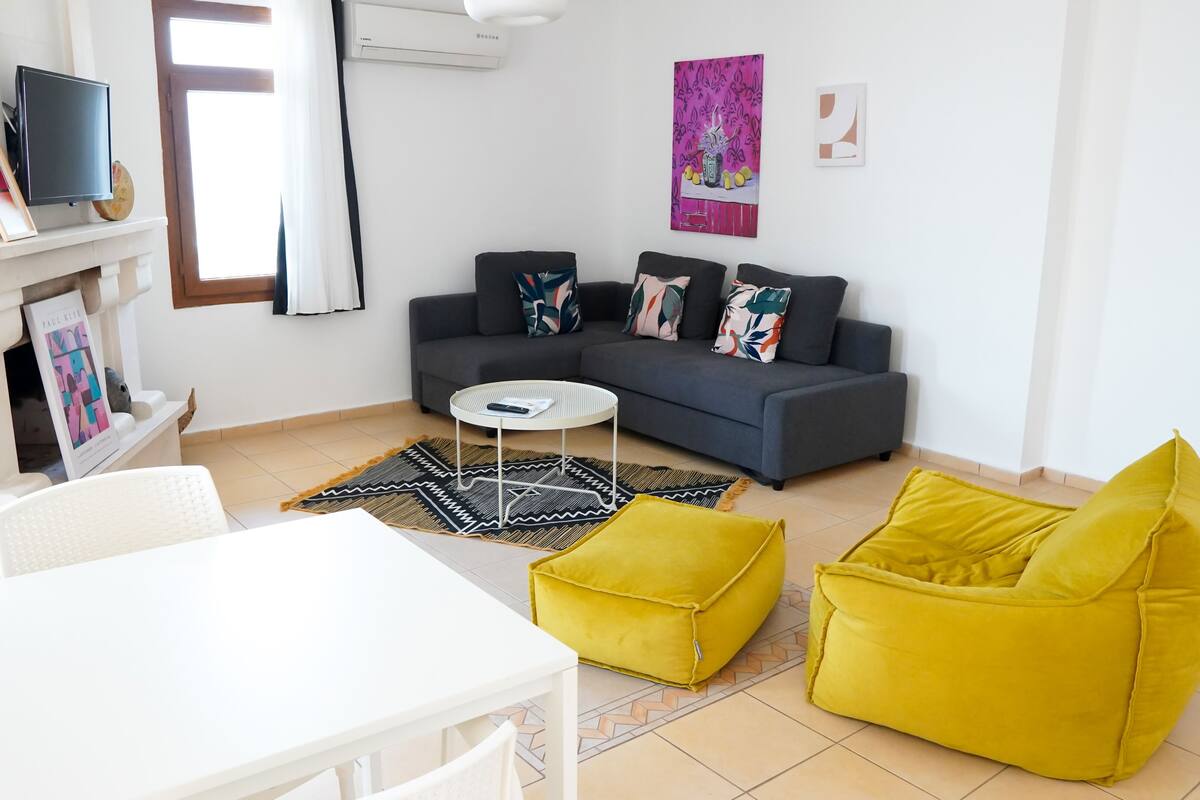 Een moderne woonkamer met een grijze bank, een geel poufje, een witte salontafel en een witte tv-meubel.