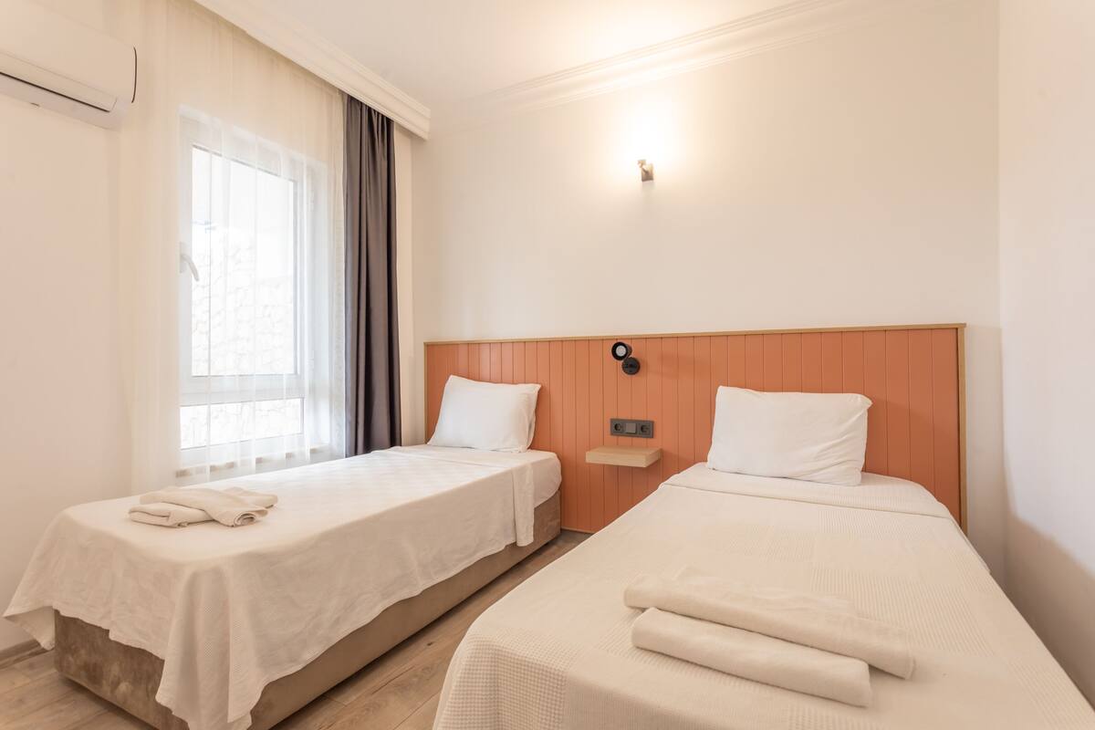 Een hotelkamer met twee enkele bedden, een klein nachtkastje, een wandlamp en een raam met gordijnen.