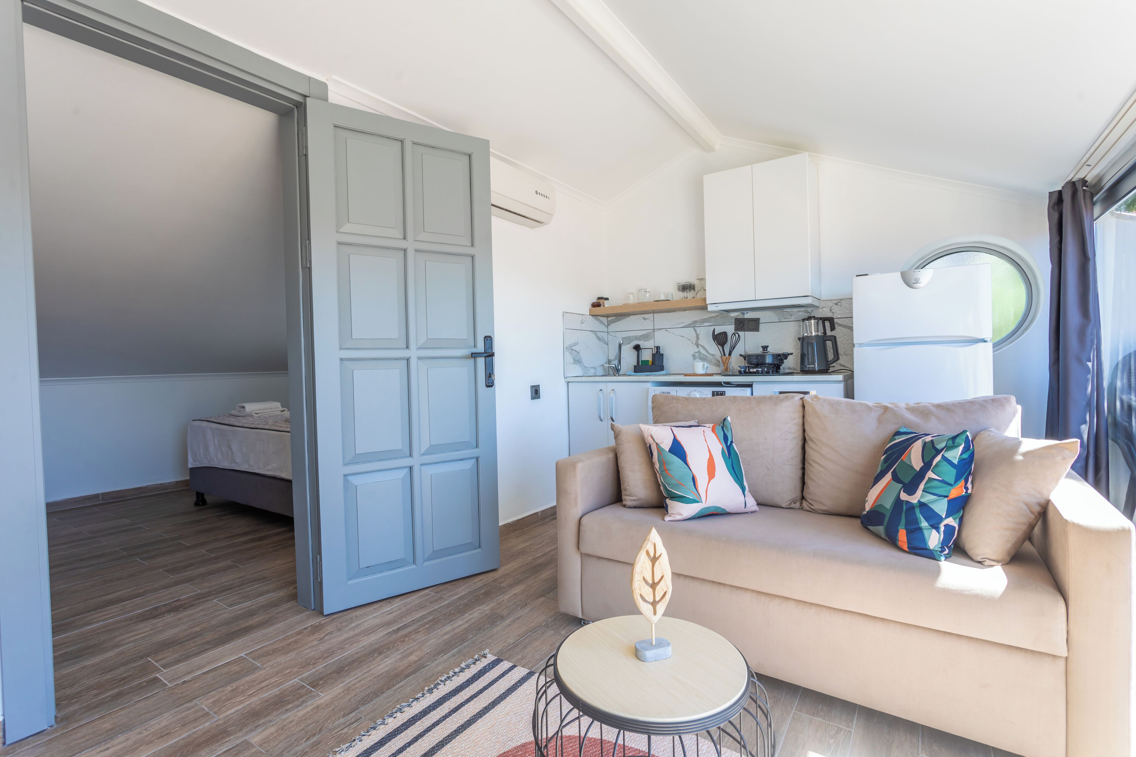 Una sala de estar moderna con un sofá, una mesita redonda y una zona de cocina con armarios y una ventana.