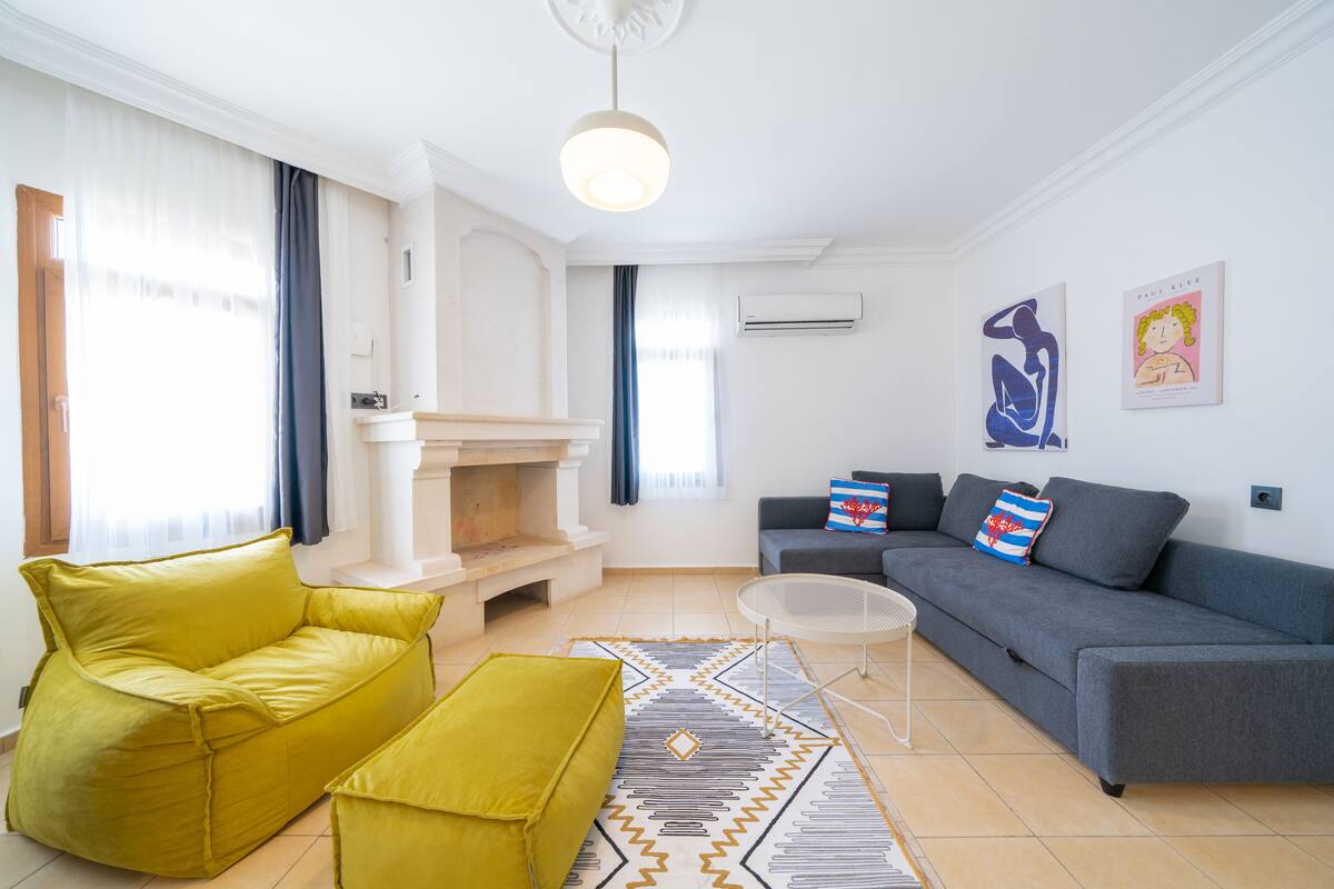 Een moderne woonkamer met een bank, poef en salontafel. Er is een open haard, een grijze bank met decoratieve kussens en kunst aan de muur.
