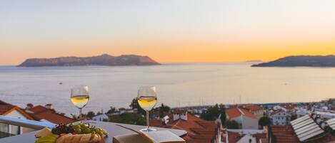A table with a book, two glasses of white wine, and a fruit platter overlooking a coastal town at sunset.