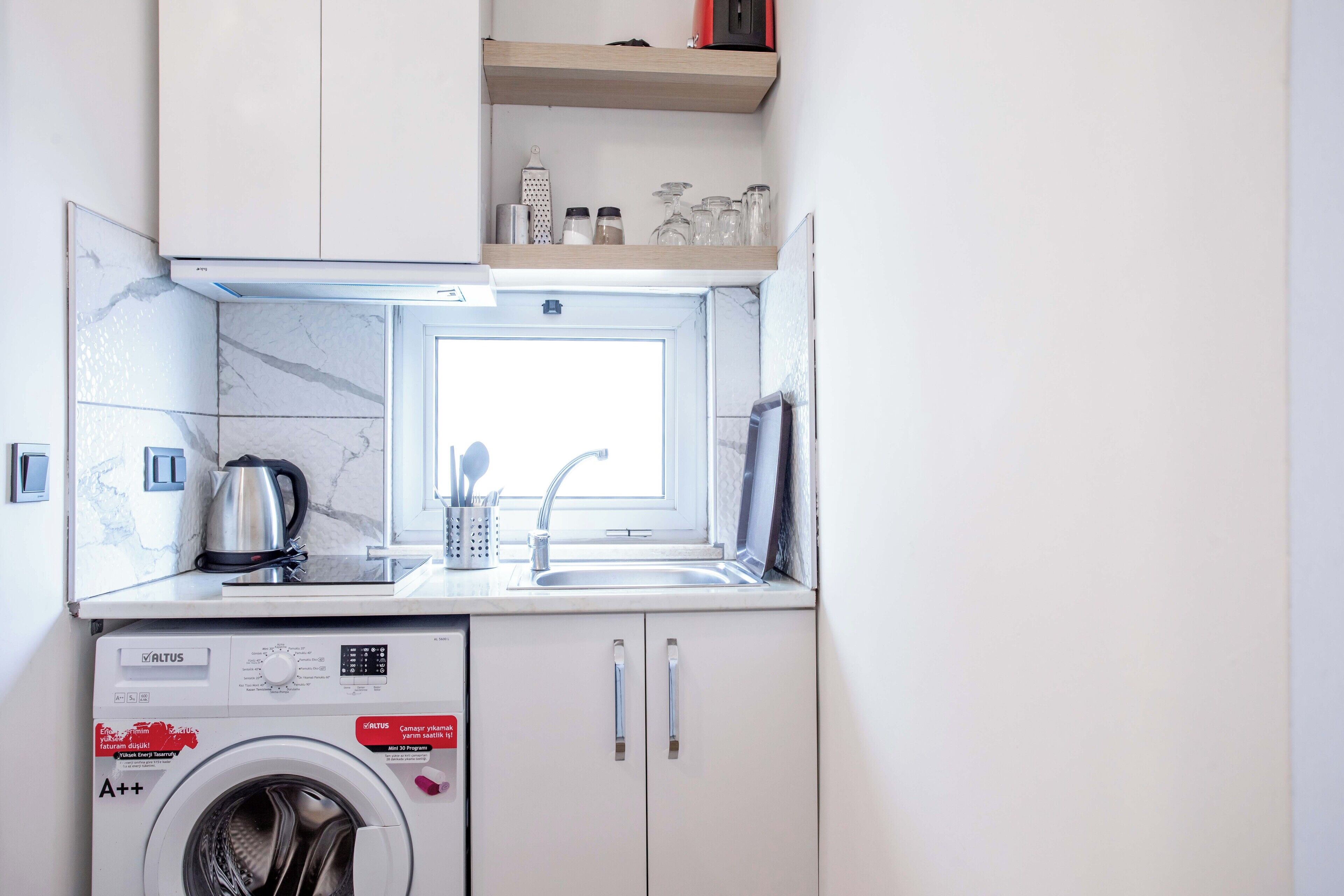 A compact kitchen with a washing machine, a kettle, and a window providing natural light.