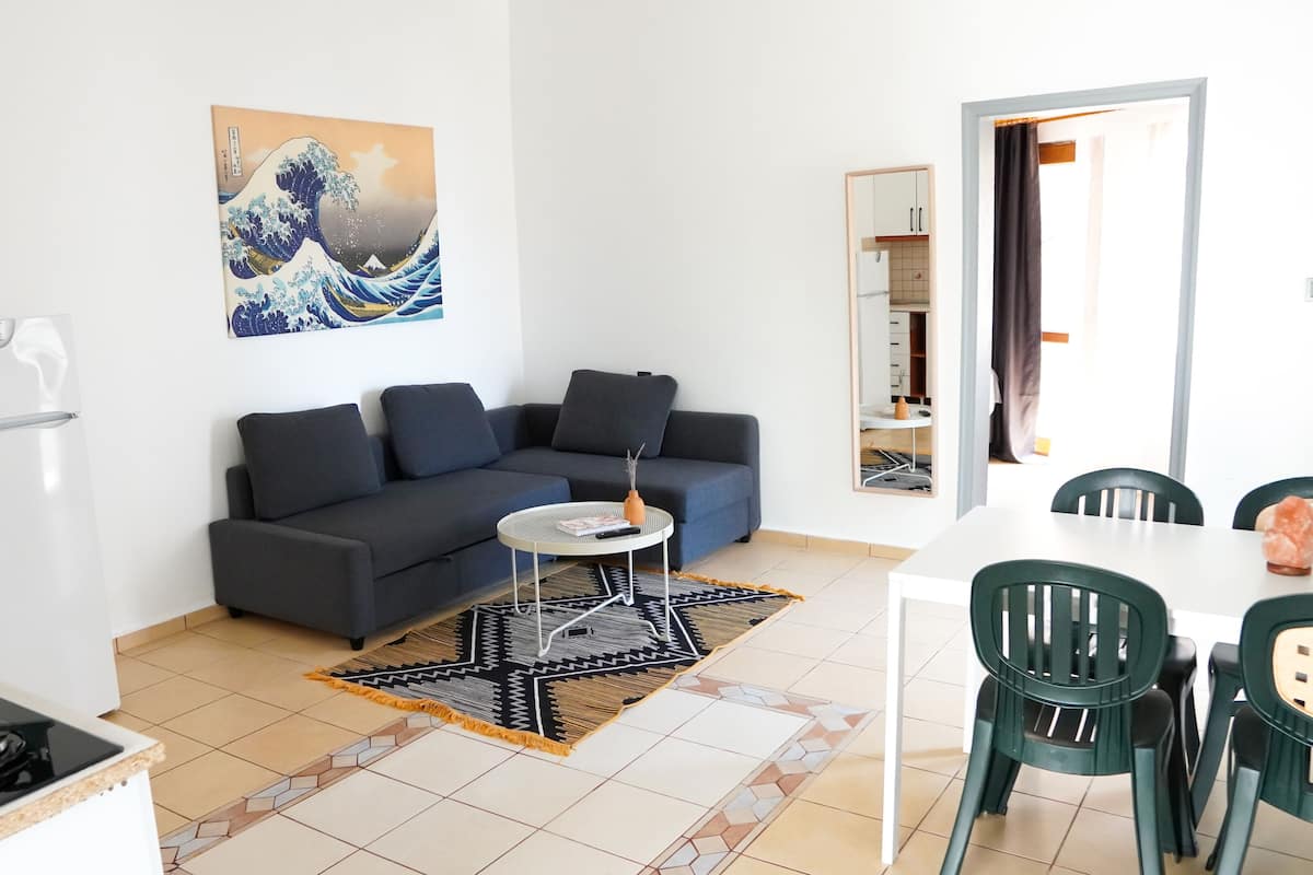 Een moderne woonkamer met een bank, een eettafel met stoelen, een keuken met een fornuis, en een groot schilderij aan de muur.