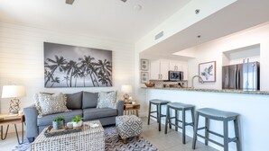 A modern living room with a gray sofa, patterned coffee table, and a large abstract painting. A kitchen area with white cabinets and a stainless steel refrigerator is visible in the background. - Seascape ~ Gulf View ~ Bunk Area (Miramar Beach)