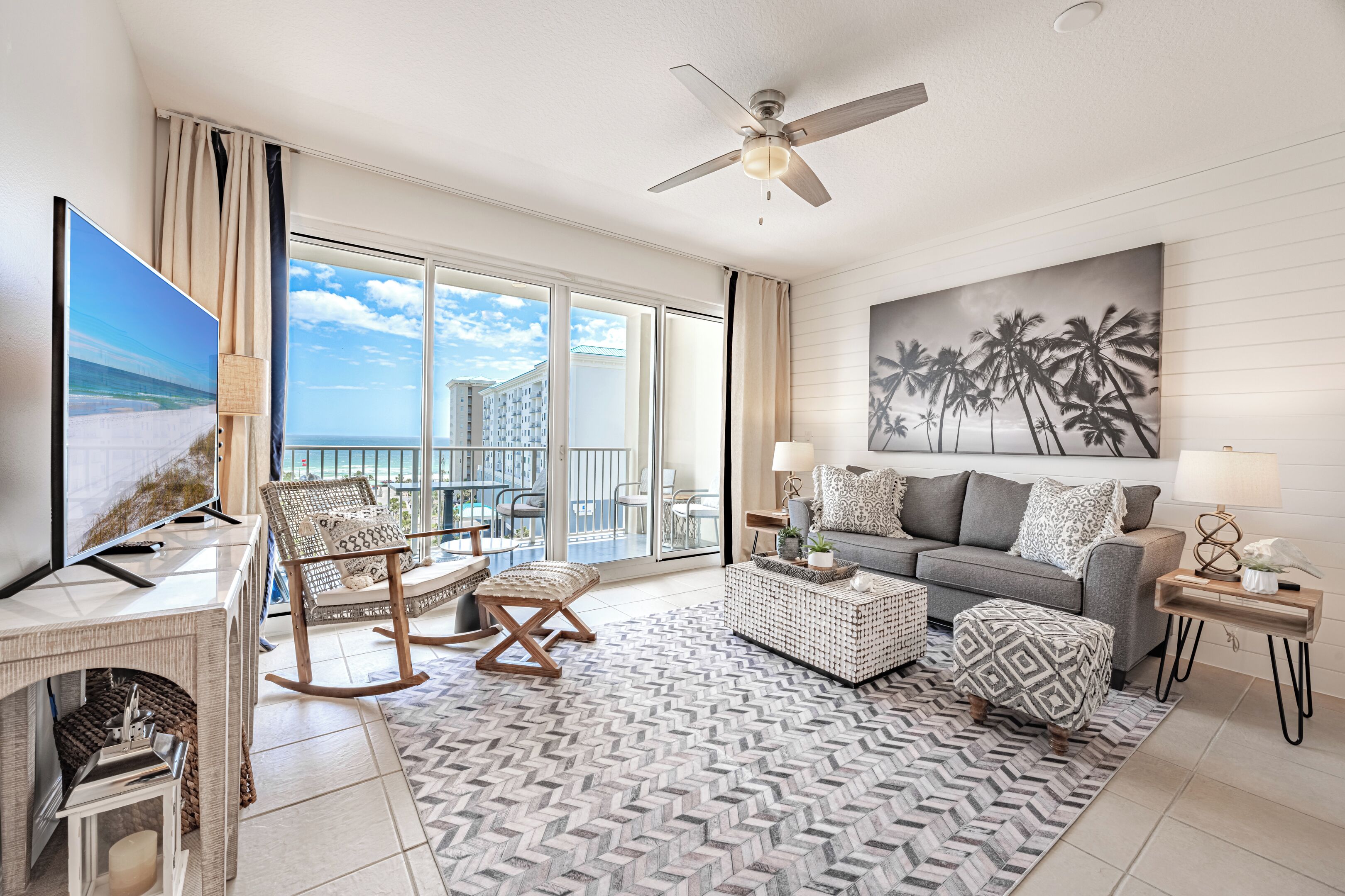 A spacious living room with a large TV, comfortable seating, a ceiling fan, and a balcony with a view of the ocean.
