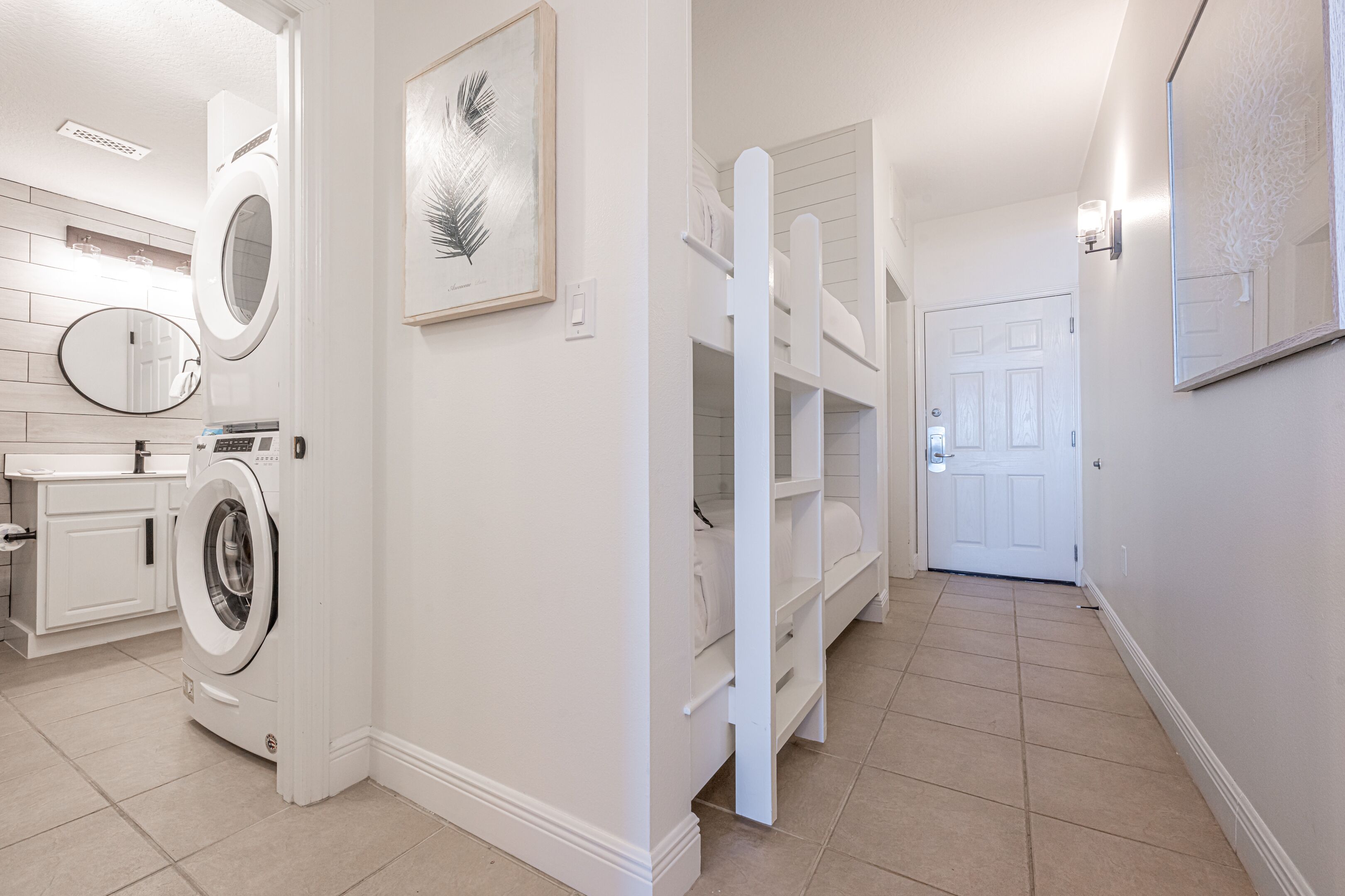 A laundry area with a washing machine, a sink, and a mirror. There are shelves with folded clothes and a door leading to another room.