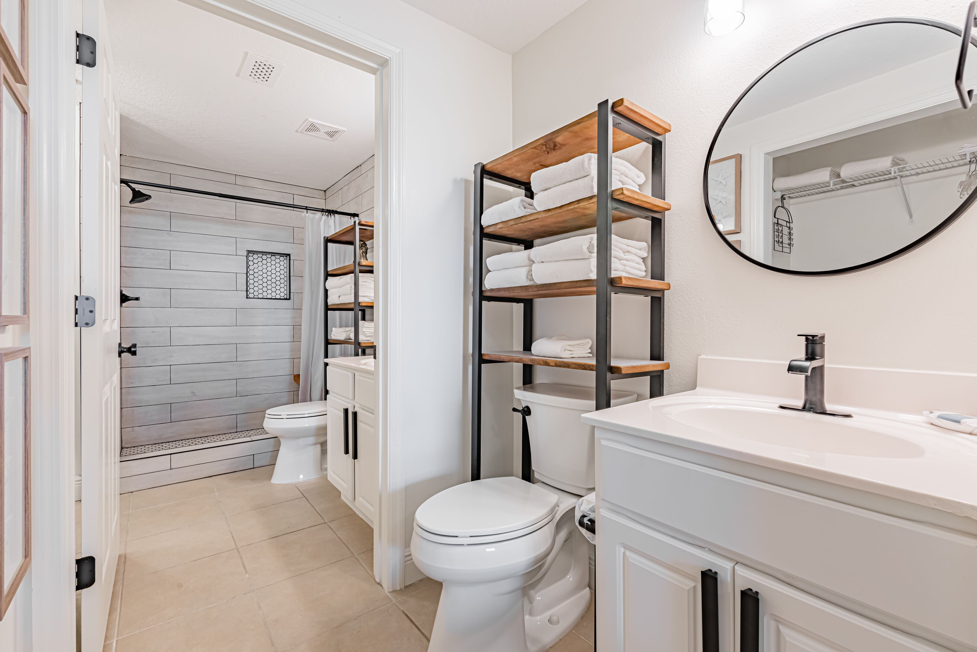 Une salle de bain avec des toilettes blanches, un lavabo et un miroir. On aperçoit une étagère avec des serviettes et une zone de douche à l’arrière-plan.