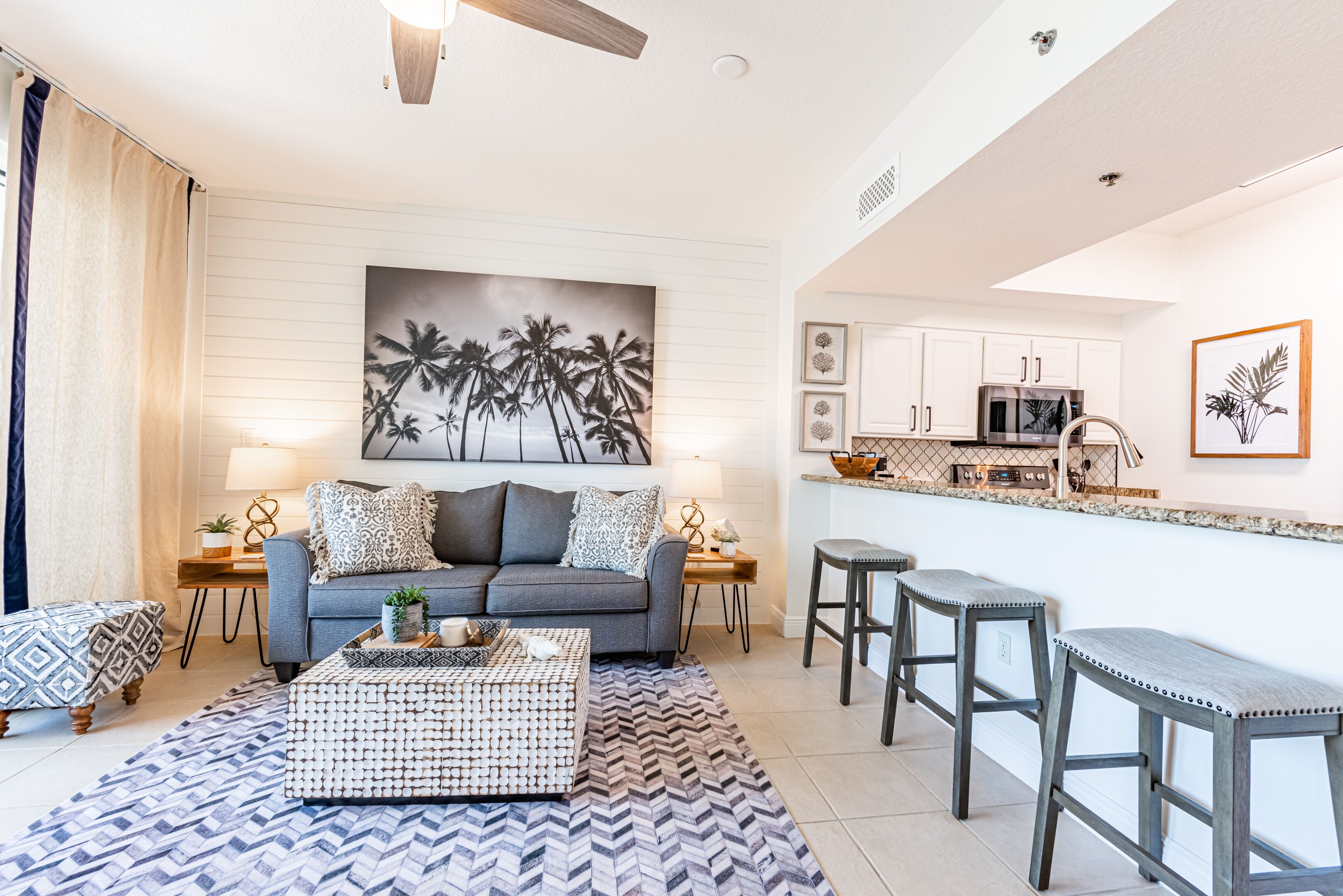 A living room with a sofa, coffee table, and patterned rug. There is a kitchen area with a microwave and bar stools.