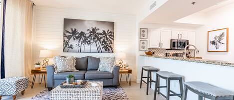 A living room with a sofa, coffee table, and patterned rug. There is a kitchen area with a microwave and bar stools.