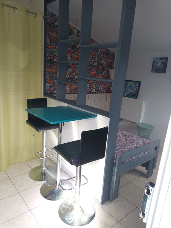 Una habitación con una cama, una mesa, dos sillas, una cortina, un estante con diseños de motivos hogareños y una pequeña mesa redonda.