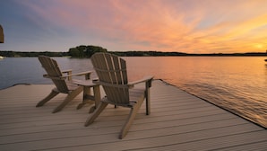 A wooden deck with two Adirondack chairs facing a calm lake at sunset.