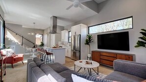 Un soggiorno moderno con un divano grigio, una poltrona rossa, una zona pranzo con sgabelli da bar, una cucina con un grande televisore e una ventola a soffitto.