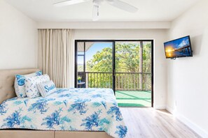 Une chambre à coucher avec un lit, une télévision, un balcon donnant sur la verdure et un ventilateur de plafond.