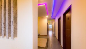 Um corredor de hotel com chão ladrilhado, uma porta de madeira à direita e uma parede com ripas de madeira verticalmente dispostas à esquerda.