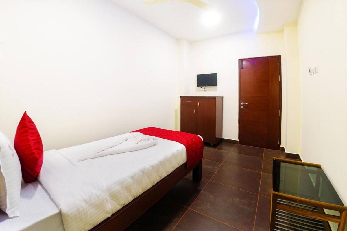 A single bed with a red and white bedspread, a wooden wardrobe, a television mounted on the wall, and a door.